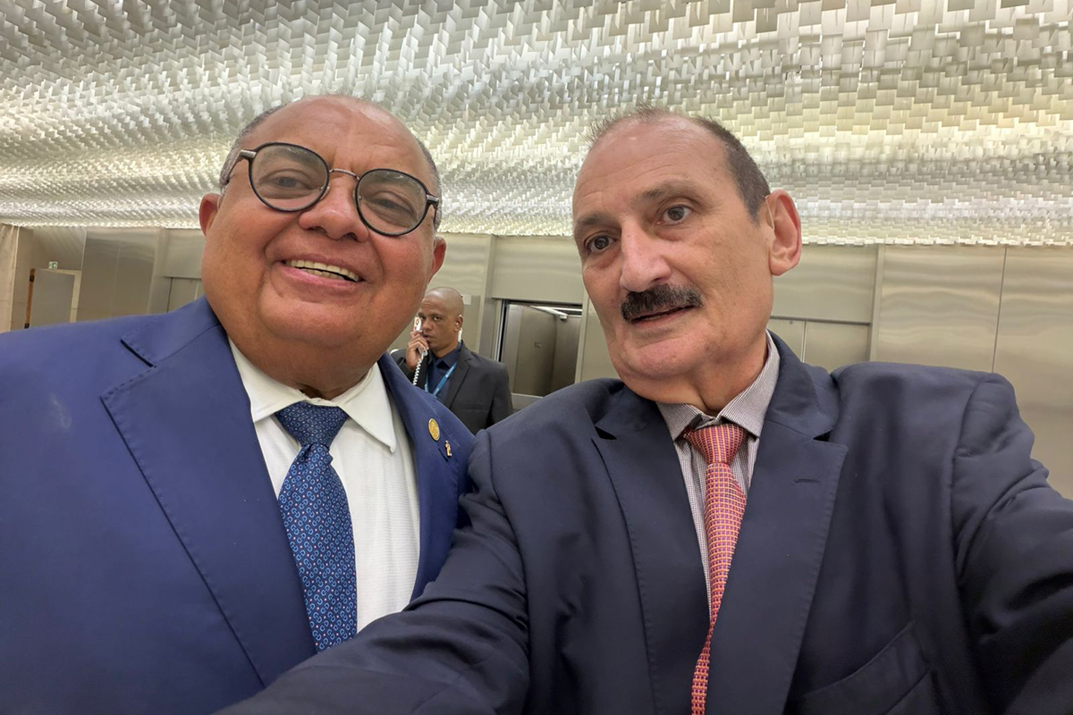 Fotografia horizontal de plano médio e corpo inteiro, tirada em ângulo frontal e nível dos olhos, com dois homens em pé em um local com teto decorativo de tiras brancas e luzes embutidas. À esquerda, um homem de pele branca, cabelos curtos e grisalhos, vestindo terno azul-marinho, camisa branca e gravata azul-clara com pequenos padrões, está de pé e sorri para a câmera. Ele usa óculos. À direita do homem, um homem de pele branca e cabelos curtos e grisalhos, vestindo terno preto, camisa branca e gravata vermelha, está de pé e olha para a câmera. Ao fundo, à esquerda, há uma pessoa de terno falando ao telefone. Mais ao fundo, em um plano de fundo borrado, há portas e paredes. A imagem é bem iluminada pela luz ambiente.