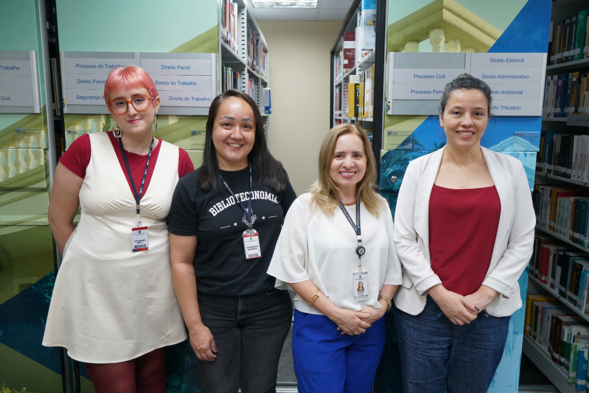 Quatro mulheres sorriem posicionadas lado a lado em um corredor de estantes da biblioteca do TRT-7. Elas vestem roupas casuais e profissionais, com crachás de identificação; a segunda mulher da esquerda para a direita usa uma camiseta preta com a inscrição "Biblioteconomia". Ao fundo, prateleiras repletas de livros jurídicos e placas indicativas de áreas do Direito compõem o cenário técnico.