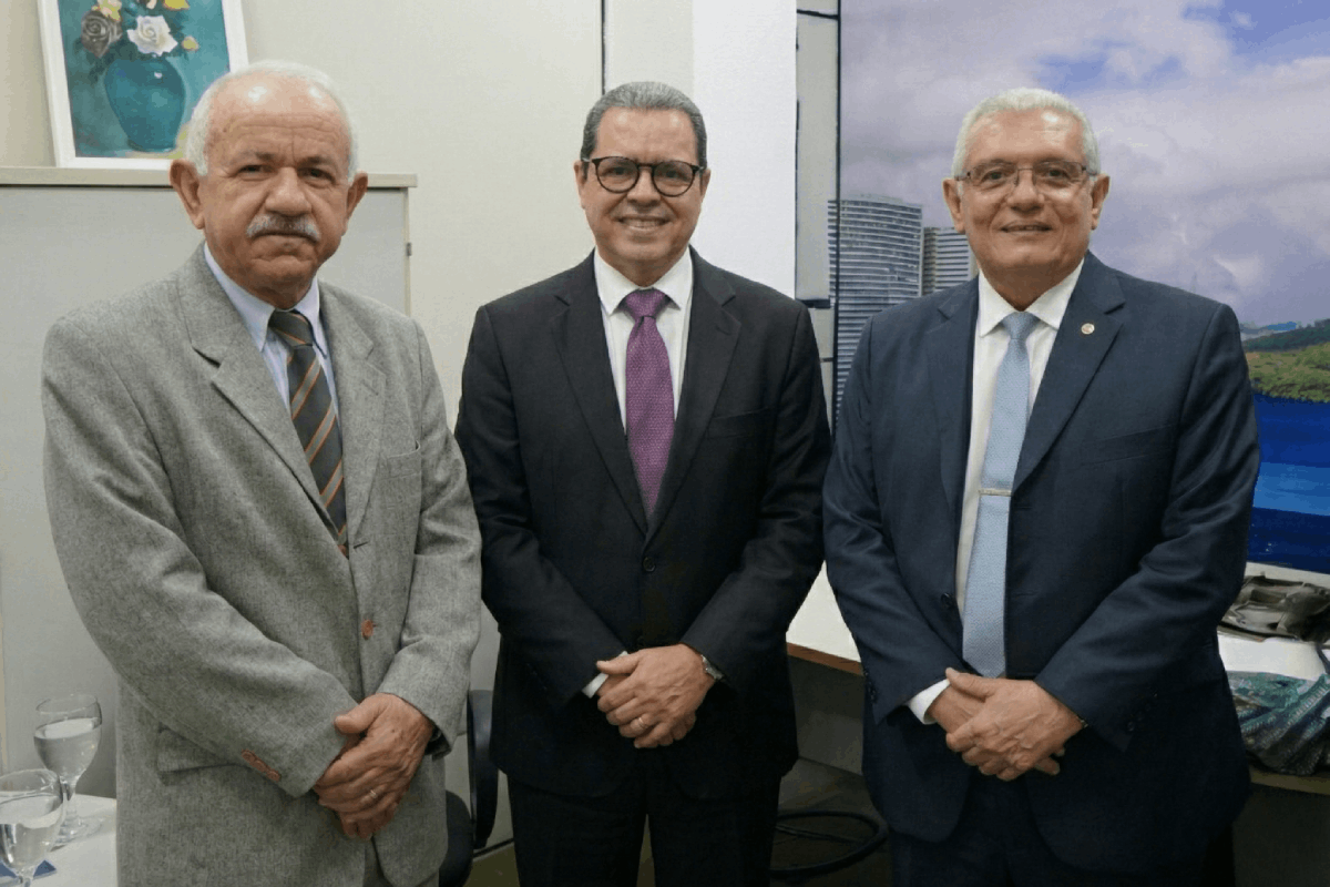 Três homens de meia-idade, em trajes formais (terno e gravata), posam lado a lado em um escritório. Eles estão de pé, com as mãos cruzadas à frente do corpo, em uma postura institucional.