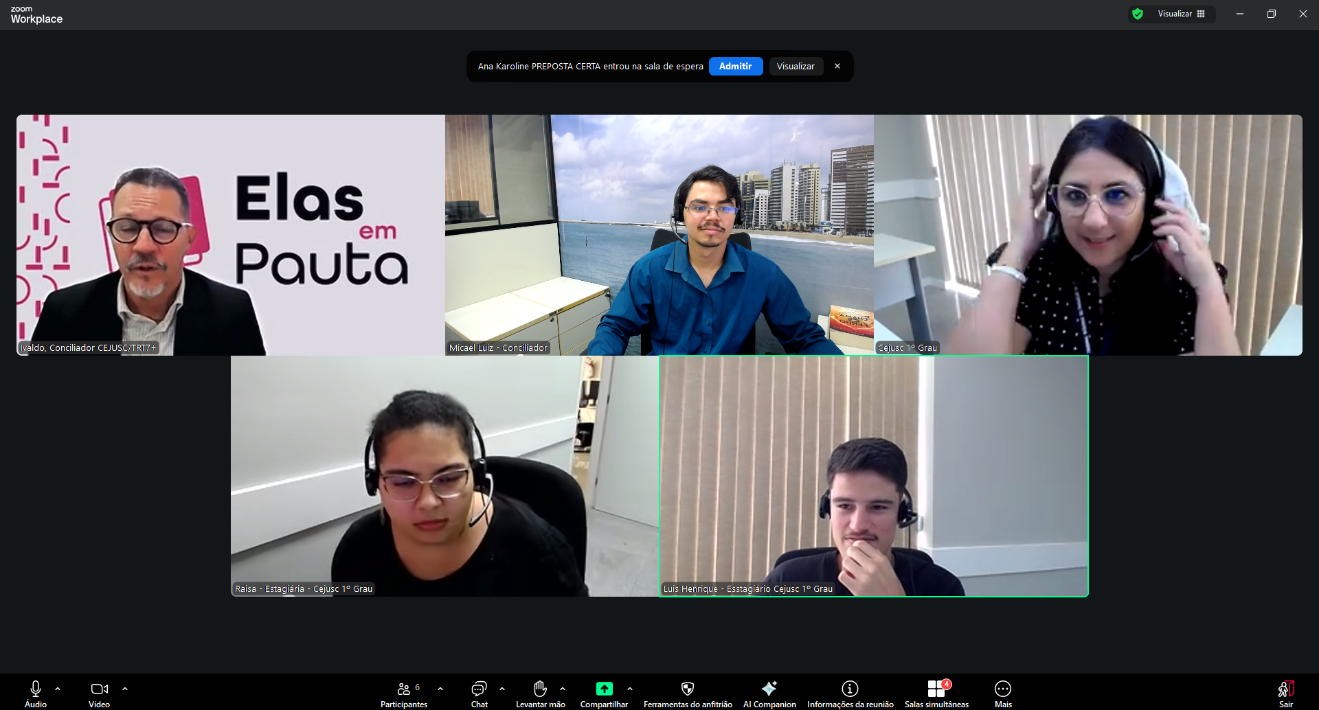 Print de tela da plataforma Zoom Workplace durante uma reunião virtual, exibindo cinco participantes em modo galeria sobre um fundo escuro. Na fileira superior, um homem com óculos usa o fundo temático "Elas em Pauta", um jovem aparece com uma vista litorânea ao fundo e uma mulher ajusta seus fones de ouvido; na fileira inferior, dois participantes utilizam fones de comunicação profissional (headsets). Na parte superior da interface, uma notificação indica que uma participante aguarda na sala de espera, enquanto a barra de ferramentas inferior mostra os ícones de controle da chamada.