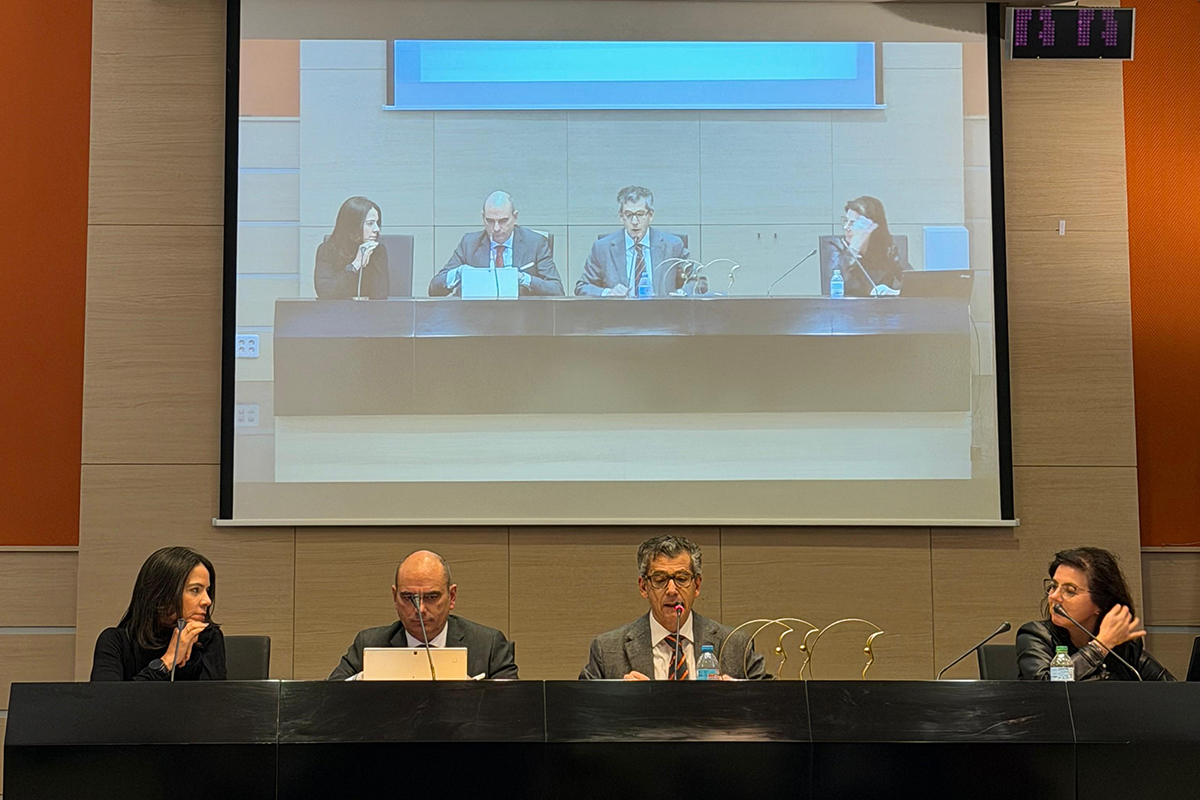 A fotografia oficial de encerramento apresenta cerca de 50 participantes reunidos em um auditório de teto arqueado e iluminação suave, posando de pé para o registro final. O grupo, composto por homens e mulheres em trajes formais de inverno, está posicionado à frente de um telão que exibe o cartaz azul do evento, emoldurado por painéis laterais em tom laranja vibrante. Em primeiro plano, as poltronas escuras do auditório aparecem em perspectiva, completando a cena que simboliza a integração das delegações no ambiente acadêmico.