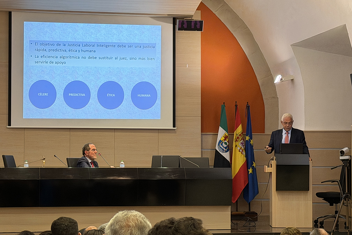  O Gemini disse A imagem mostra uma palestra em um auditório moderno, onde um homem grisalho discursa em um púlpito ao lado das bandeiras da Extremadura, Espanha e União Europeia. Ao fundo, uma projeção em espanhol destaca os pilares da "Justiça Laboral Inteligente": célere, preditiva, ética e humana. Em uma mesa comprida à esquerda, outro homem acompanha a apresentação, enquanto o topo das cabeças da plateia aparece em primeiro plano.