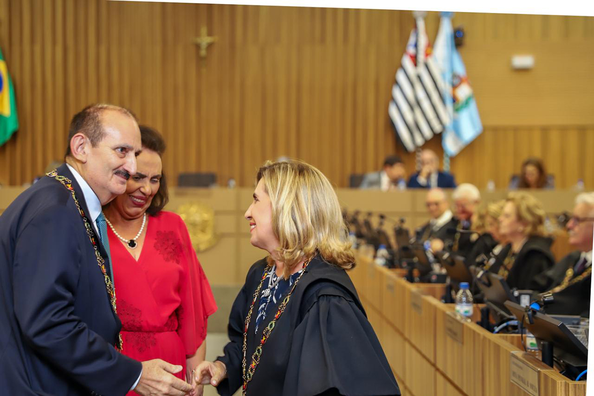 Em um ambiente solene, um homem de terno e uma mulher de toga preta, ambos usando colares de honraria, trocam um aperto de mão e sorrisos. Entre eles, uma mulher em um vestido vermelho vibrante observa o gesto com expressão alegre, enquanto ao fundo vê-se uma bancada com outras autoridades sentadas. A cena é emoldurada por painéis de madeira e pelas bandeiras de São Paulo e do Rio de Janeiro