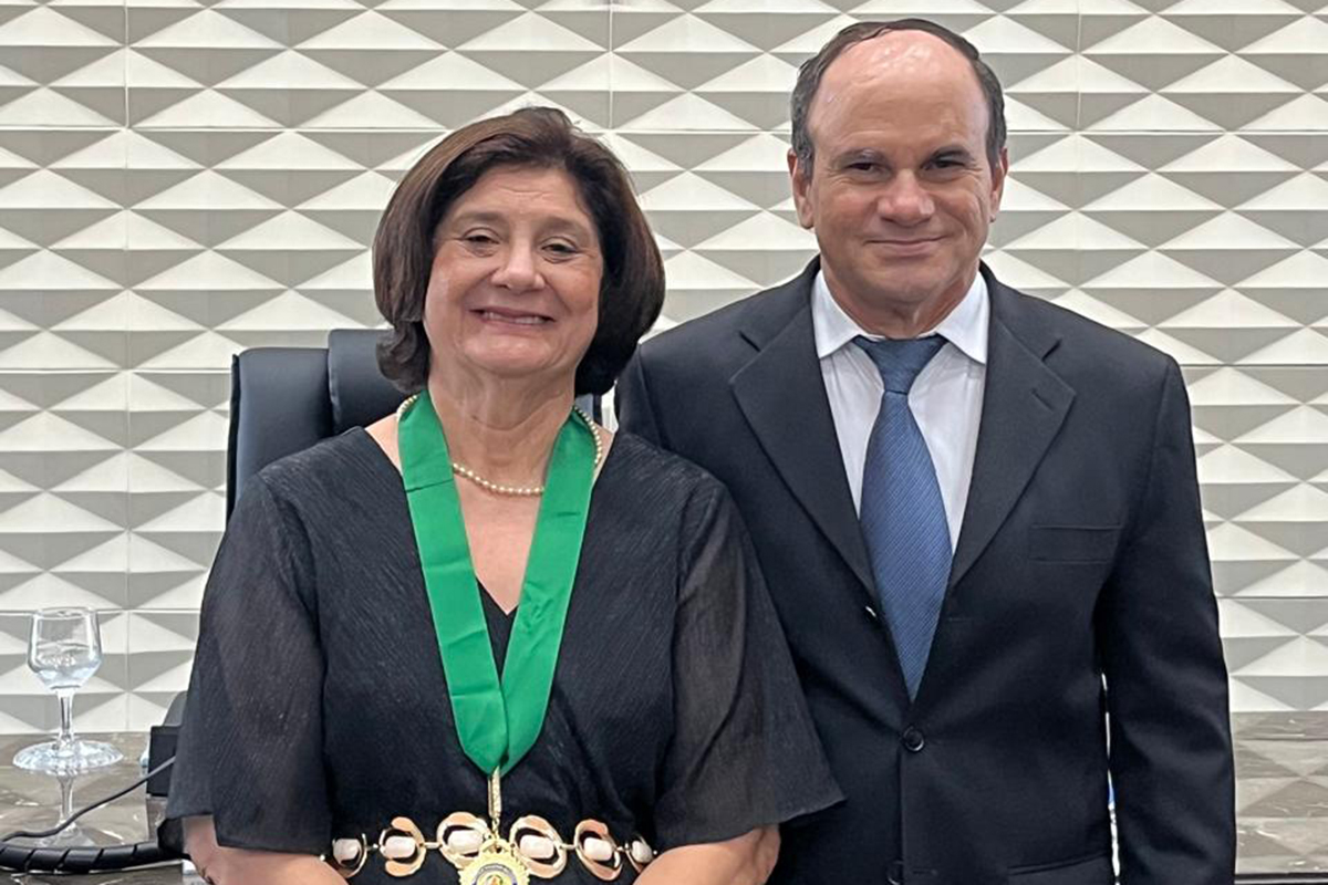 Retrato em plano médio de uma mulher e um homem sorridentes, em pé, lado a lado, em um ambiente interno. A mulher, à esquerda, usa um vestido preto, um colar de pérolas, uma fita verde no pescoço e um cinto com detalhes. O homem veste terno e gravata. Ao fundo, há uma parede com padrão geométrico e um crucifixo pendurado no centro.