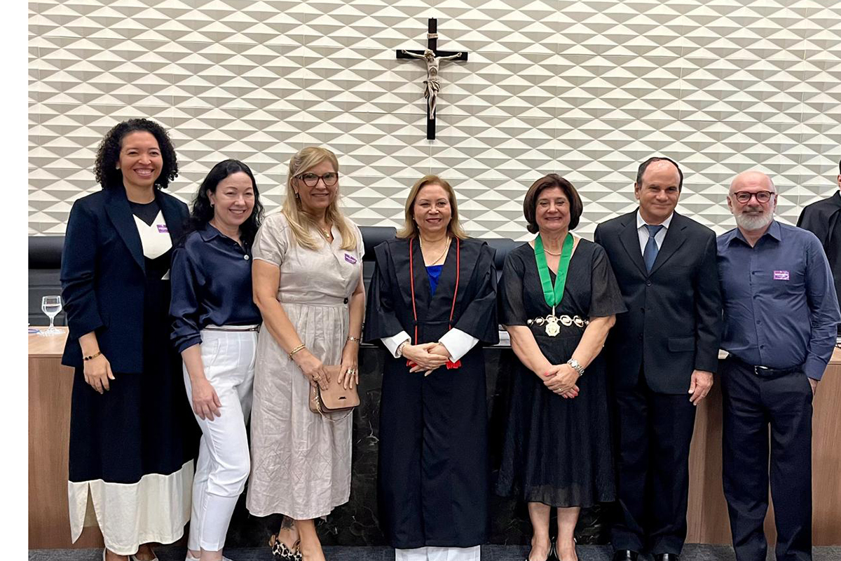 Sete pessoas posam para foto em pé, lado a lado, em um plenário com parede geométrica e um crucifixo ao centro. No meio, uma mulher veste toga preta com cordões vermelhos e, à sua esquerda, outra mulher usa um vestido preto com fita verde e medalha. Os demais homens e mulheres vestem trajes sociais variados, como ternos, camisas e vestidos casuais, todos sorrindo para a câmera.
