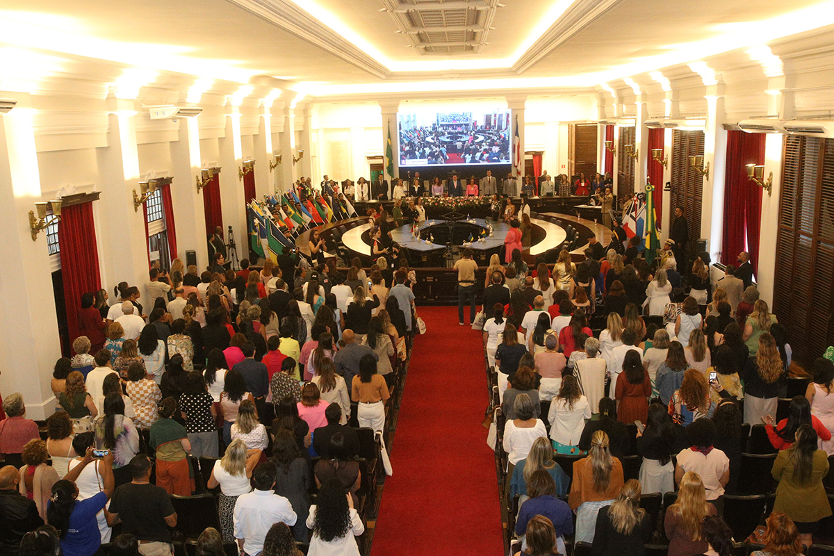 Plenário do Fórum Ruy Barbosa lotado com a plateia majoritariamente feminina em pé, voltada para a mesa de honra ao fundo. Um tapete vermelho central divide as fileiras de poltronas e conduz o olhar para o telão e as bandeiras coloridas que decoram o ambiente solene. A iluminação é clara e destaca a arquitetura clássica do salão com grandes janelas de madeira e detalhes no teto.
