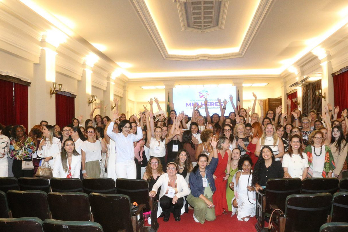 Um numeroso grupo de mulheres, composto por magistradas e servidoras, posa para foto no salão nobre do Fórum Ruy Barbosa, em Salvador. Muitas estão com os braços erguidos em sinal de celebração, preenchendo o espaço entre as fileiras de poltronas escuras e o telão ao fundo. O ambiente possui arquitetura clássica, com teto ornamentado, colunas claras e grandes janelas com cortinas vermelhas.