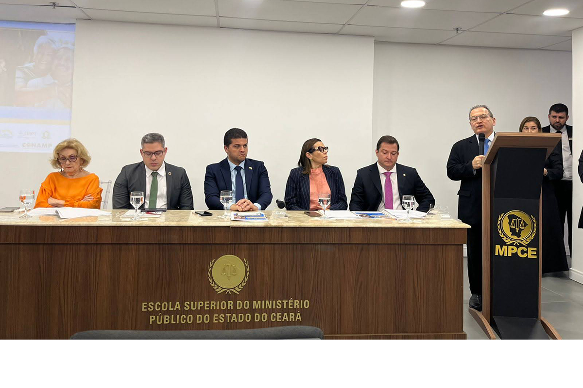 O Gemini disse Esta imagem captura uma cerimônia formal em um auditório da Escola Superior do Ministério Público do Estado do Ceará (ESMP/CE), apresentando um grupo de autoridades alinhadas e de pé no centro da sala, enquanto a plateia ocupa assentos em primeiro plano e ao fundo. No centro do grupo, uma mulher de blusa laranja vibrante e calças com padrão de folhagem colorida segura um documento, flanqueada por homens e mulheres vestidos com trajes sociais, como ternos azuis e cinzas, blazers rosa e preto, e vestidos de cores sólidas. Atrás deles, há uma projeção de slides em uma tela branca, e à direita, um púlpito com a inscrição "MPCE" e uma televisão transmitindo o evento, que exibe o título "Lançamento da Campanha: Justiça que cuida". O ambiente é bem iluminado por luzes embutidas no teto de gesso, com saídas de ar condicionado visíveis, e o chão é revestido com piso frio cinza. A mesa principal ao fundo exibe o brasão da ESMP/CE e a inscrição "ESCOLA SUPERIOR DO MINISTÉRIO PÚBLICO DO ESTADO DO CEARÁ".