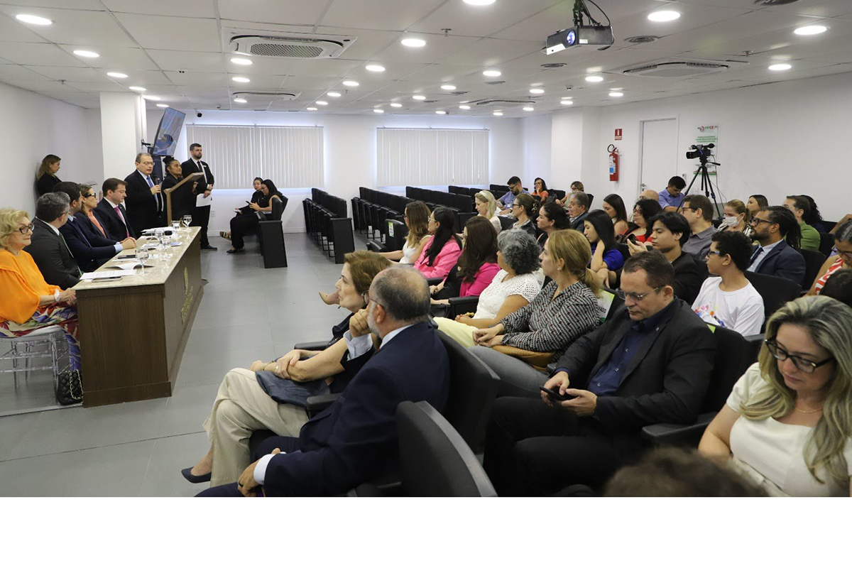 As imagens mostram o lançamento de uma campanha institucional em um auditório climatizado e bem iluminado. Na primeira imagem, um grupo de 11 pessoas posa em pé e sorrindo, segurando uma placa que celebra a "Justiça que cuida".  Detalhes Visuais: O grupo na primeira imagem está elegantemente vestido e organizado em frente a um pano de fundo que exibe o nome da campanha e logomarcas oficiais, incluindo a do Governo do Ceará. Na segunda imagem, vê-se o auditório repleto de pessoas sentadas em cadeiras de estofado escuro, assistindo a uma palestra.  Cenário e Texto: O auditório da segunda imagem tem um palco ao fundo onde um palestrante fala ao microfone. Uma câmera de vídeo profissional em um tripé está posicionada à direita para registrar o evento.