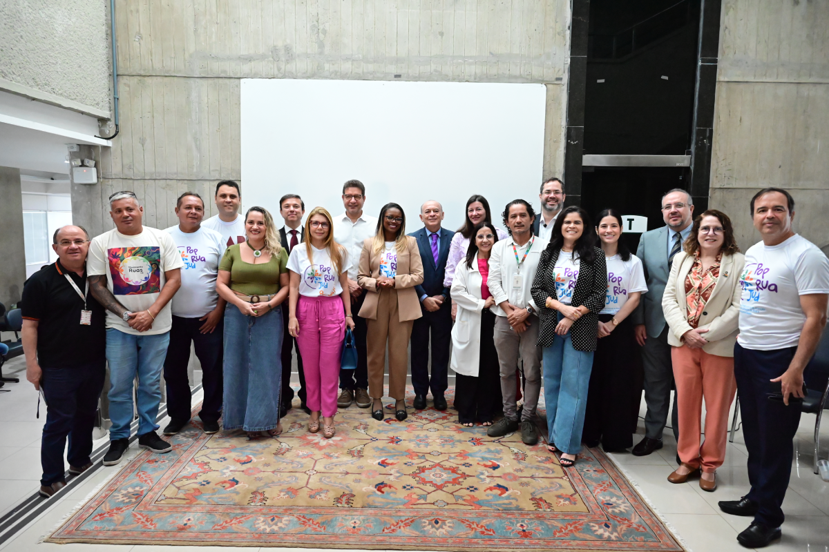 Um grupo diversificado de aproximadamente vinte pessoas posa sorridente sobre um grande tapete persa em um ambiente interno de concreto aparente. Muitos vestem a camiseta branca oficial do evento com o logotipo colorido "PopRuaJud", alternando com outros em trajes formais como paletós e blazers. Ao fundo, uma parede clara com um painel retangular branco centraliza a composição do registro coletivo.