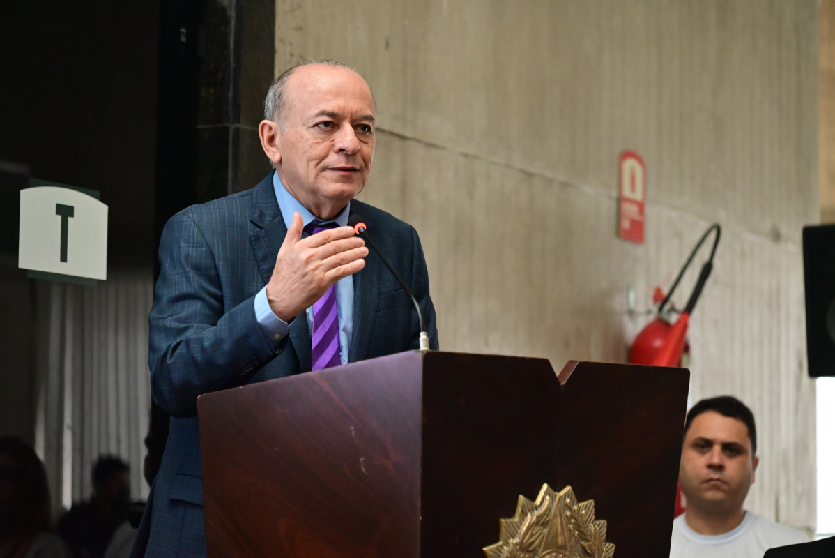 Homem de pele clara e cabelos ralos discursa em um púlpito de madeira escura adornado com o brasão da República. Ele veste terno xadrez azul-marinho, camisa azul-clara e gravata roxa listrada, gesticulando com a mão direita próxima ao microfone. Ao fundo, uma parede de concreto aparente exibe um extintor de incêndio, enquanto um homem observa a cena à direita.