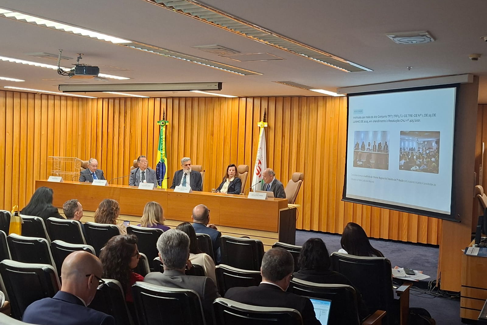 Fotografia de um auditório onde um homem de terno preto, sentado à direita de uma bancada de madeira, discursa. Atrás dele, mais quatro pessoas de traje formal estão sentadas na bancada, com as bandeiras do Brasil e de uma organização oficial ao fundo. À direita, um telão projeta um slide com texto, e a audiência composta por dezenas de pessoas sentadas em poltronas azuis observa a apresentação.