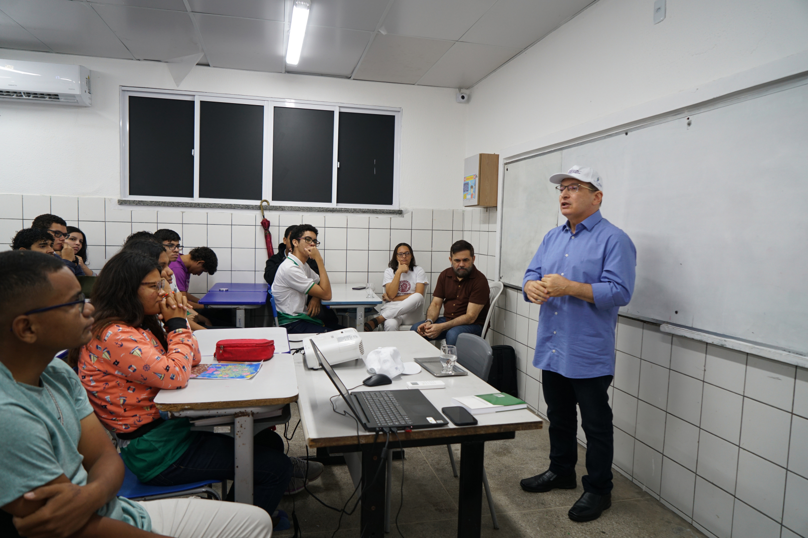 Um homem de camisa azul e boné branco fala em pé ao lado de uma lousa branca, enquanto um grupo de jovens estudantes o observa atentamente sentados em suas bancas. No primeiro plano, uma mesa branca apoia um notebook aberto, um projetor e um celular, com as paredes da sala revestidas por azulejos brancos na parte inferior. Ao fundo, outros adultos acompanham a apresentação em uma sala de aula iluminada por luzes fluorescentes.