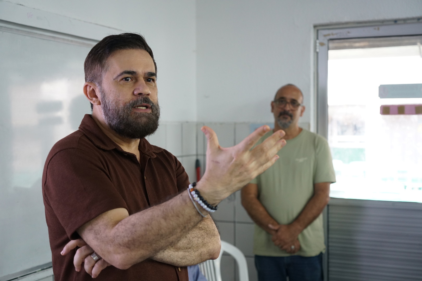 Uma foto de médio plano, ligeiramente de baixo, captura o orador de barba cerrada, vestindo uma camisa de botões marrom, no canto superior esquerdo. Ele tem o braço direito cruzado sobre o peito e o esquerdo levantado e estendido para a direita com a palma da mão voltada para cima em um gesto de oratória. Ao fundo, no canto inferior direito, um homem careca com óculos e barba vestindo uma camiseta verde-clara olha para ele, com as mãos cruzadas à frente. O ambiente é um ambiente interno utilitário com azulejos de parede brancos, uma porta de alumínio e vidro à direita e um quadro branco esmaecido à esquerda.