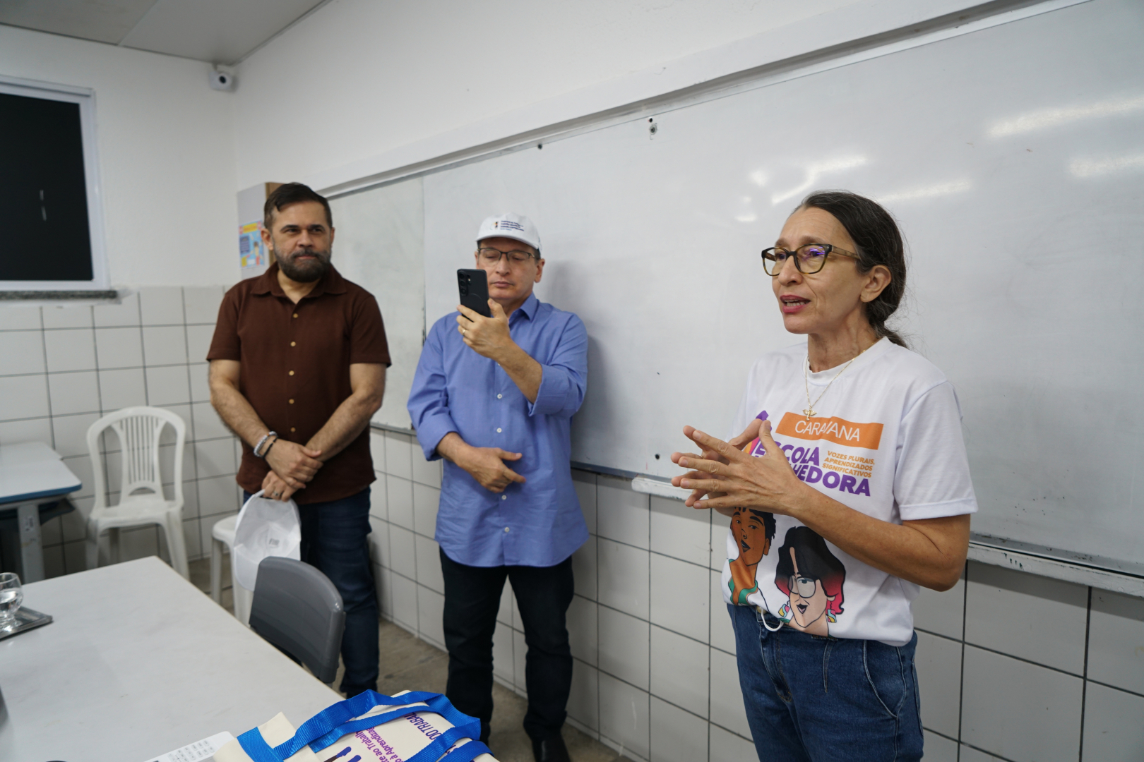  O Gemini disse Uma mulher de óculos e camiseta branca da "Caravana Escola Acolhedora" fala e gesticula com as mãos em primeiro plano. Atrás dela, um homem de camisa azul registra o momento com um celular, enquanto outro homem de camisa marrom observa a cena com as mãos cruzadas. O grupo está em uma sala de aula com paredes de azulejos brancos e um grande quadro branco ao fundo.