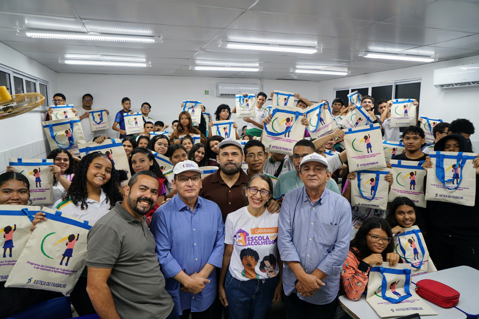 Uma fotografia colorida captura dezenas de estudantes e alguns adultos posando em pé em uma sala de aula de paredes brancas, segurando sacolas de pano brancas com alças azuis e o logotipo da Justiça do Trabalho. No primeiro plano, uma mulher com uma camiseta branca que diz "ESCOLAS ACOLHEDORAS" e dois homens vestindo camisas de botão (uma azul e outra cinza) sorriem para a câmera. O teto é revestido por painéis brancos e luzes fluorescentes embutidas.