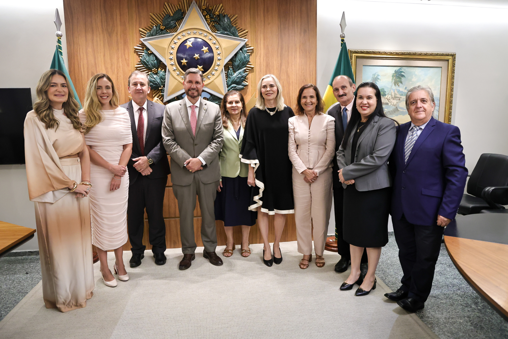 Dez autoridades, seis mulheres e quatro homens, posam para uma foto oficial em uma sala com painéis de madeira e o Brasão de Armas do Brasil ao centro. Eles vestem trajes formais, como ternos, vestidos e conjuntos de alfaiataria, posicionados entre bandeiras oficiais e uma pintura de paisagem litorânea ao fundo. O grupo sorri para a câmera, mantendo as mãos cruzadas à frente ou ao lado do corpo sobre um tapete claro.