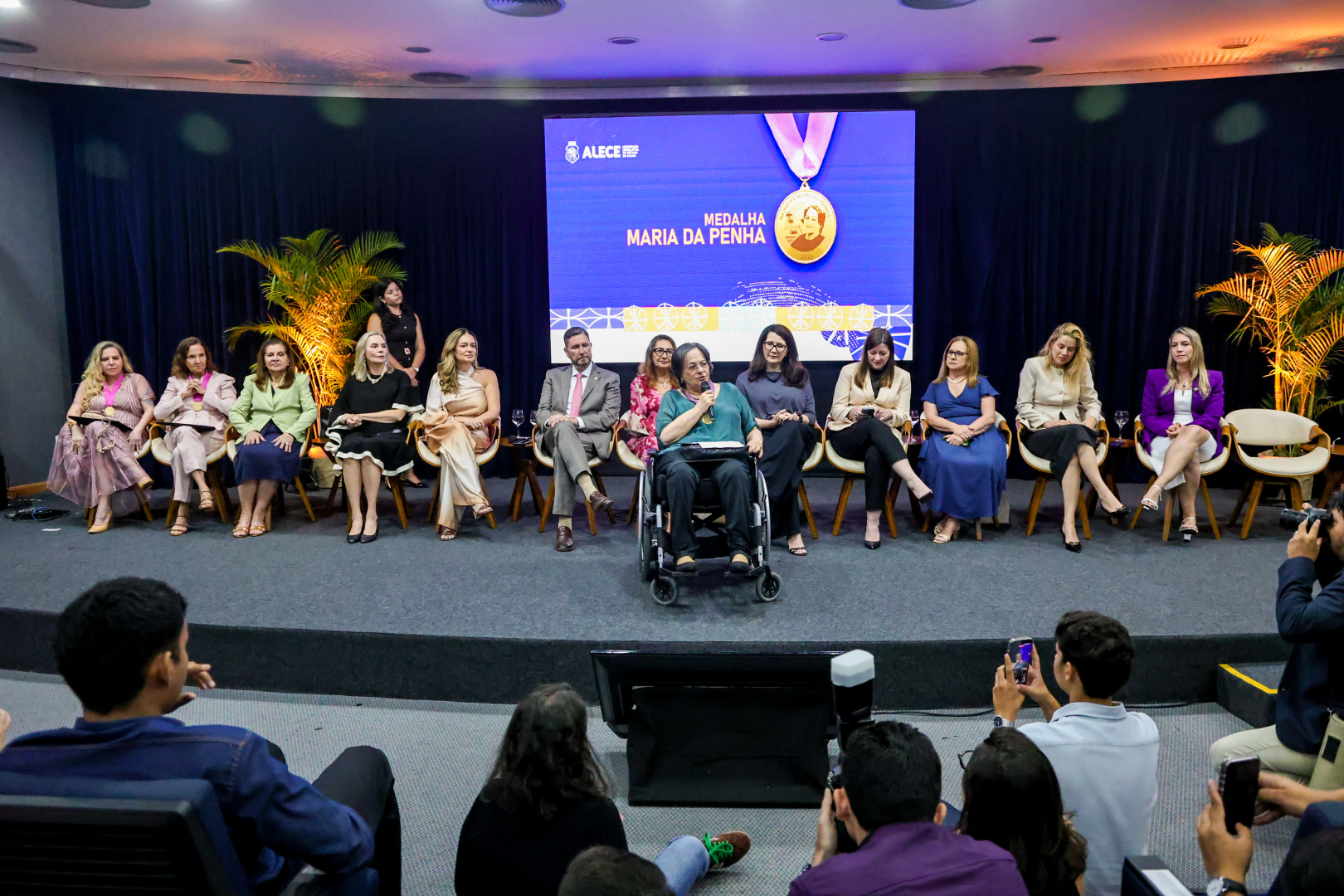 Em um auditório, Maria da Penha fala ao microfone em sua cadeira de rodas, posicionada à frente de uma longa fila de autoridades sentadas em um palco com cortinas azuis. Ao fundo, um telão exibe a imagem de uma medalha de ouro sob o título "Medalha Maria da Penha". Na plateia, em primeiro plano, diversas pessoas registram o momento com celulares e câmeras fotográficas.