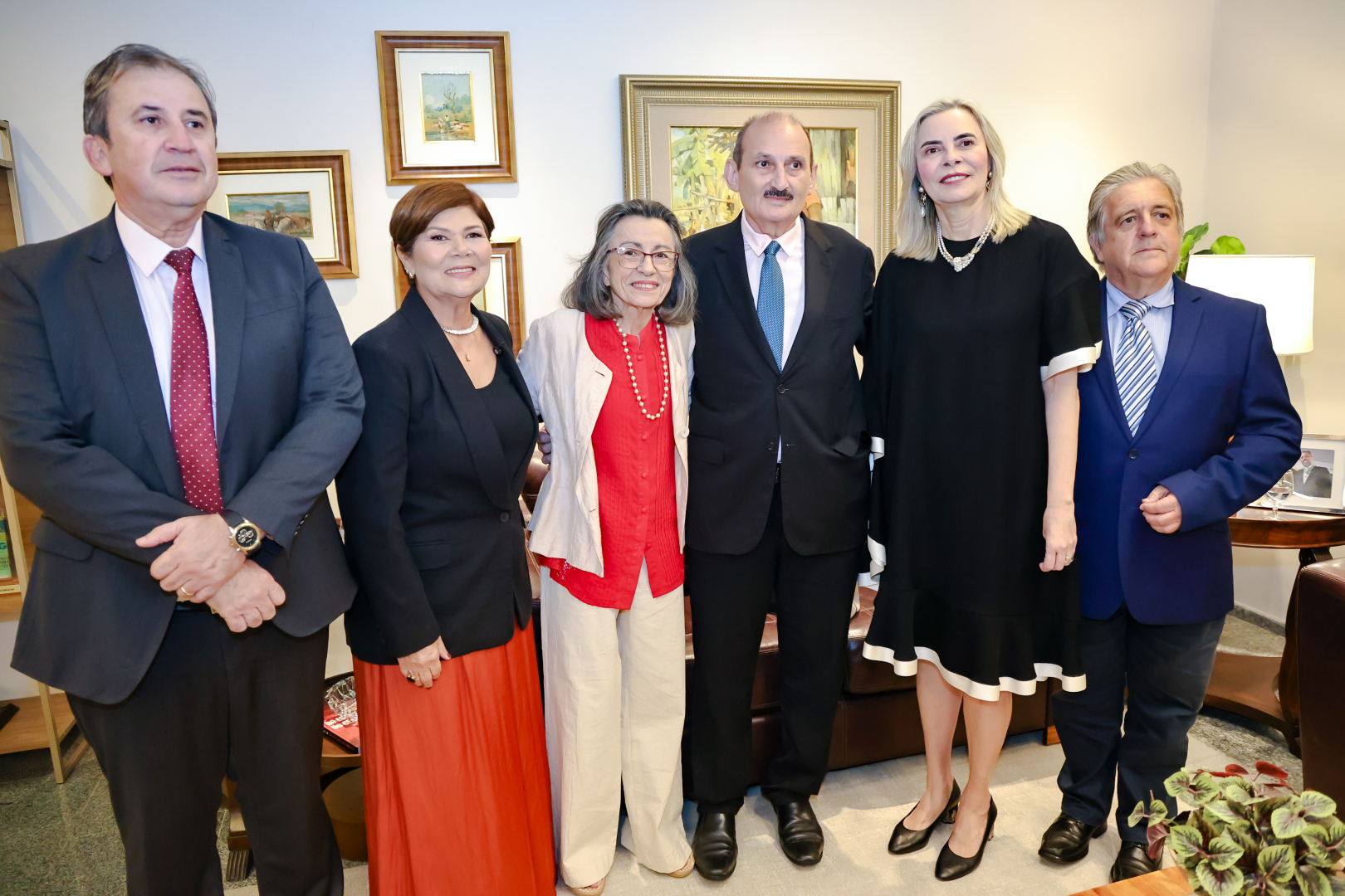 Três mulheres e três homens posam em pé, lado a lado, em uma sala decorada com quadros em molduras clássicas. Eles vestem roupas sociais em tons de preto, azul, branco e vermelho, destacando-se uma mulher ao centro com blusa vermelha e colar de pérolas. O grupo sorri cordialmente para a câmera, posicionados à frente de um sofá de couro marrom e móveis de apoio.