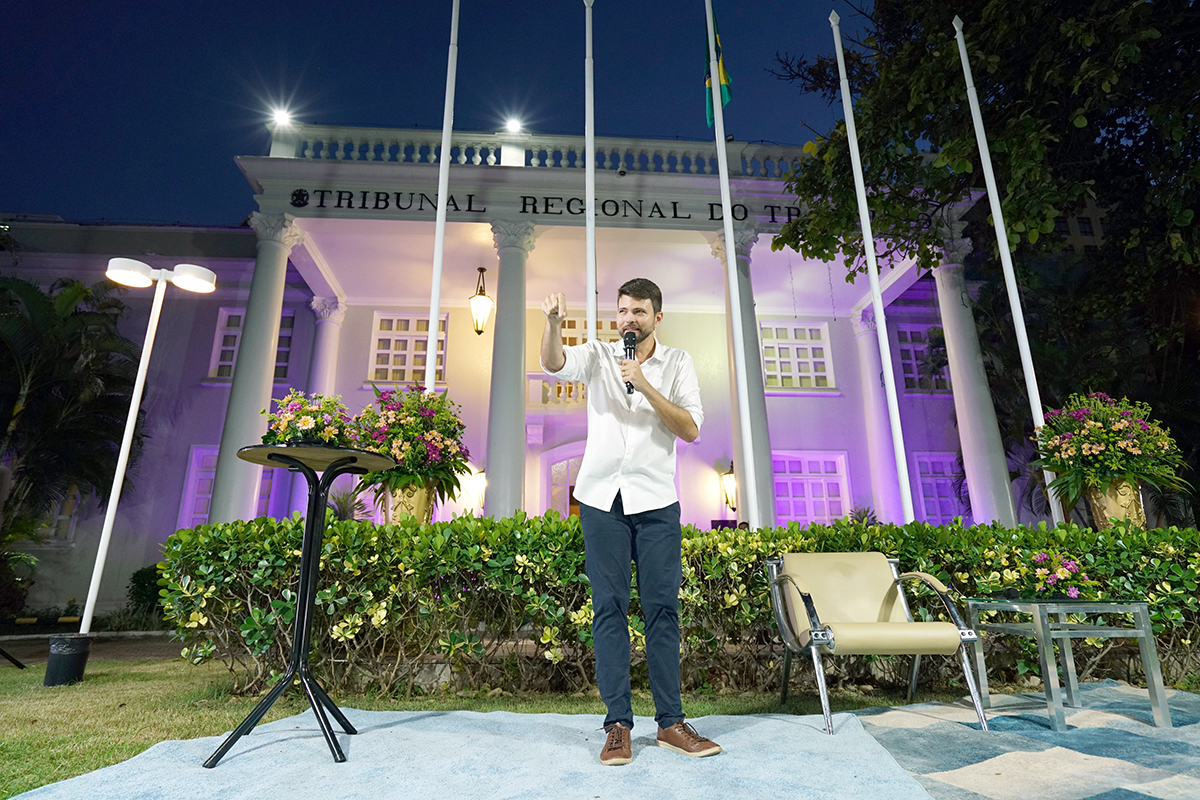 Um homem jovem, de camisa branca e calça escura, fala ao microfone em um palco ao ar livre à noite. Ao fundo, a fachada iluminada do Tribunal Regional do Trabalho, com colunas brancas e bandeiras. O ambiente é decorado com arranjos de flores coloridas e poltronas modernas sobre um tapete claro.