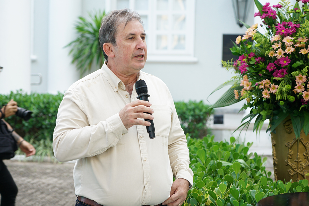 Plano médio de um homem de meia-idade, com cabelos grisalhos e camisa social bege, falando ao microfone. Ele está em um ambiente externo com vegetação verde ao fundo. À direita, destaca-se um grande arranjo de flores em tons de rosa e amarelo em um vaso ornamental dourado.