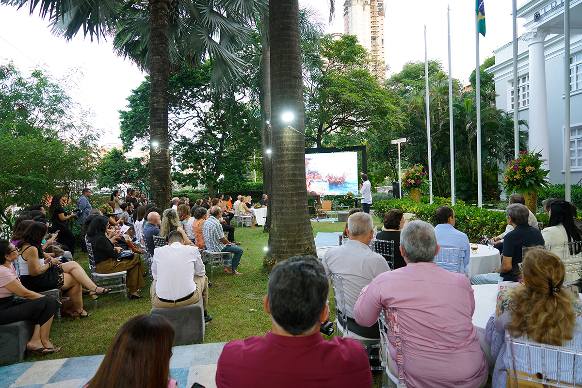 Vista panorâmica de um evento ao ar livre em um jardim com palmeiras imperiais. O público está sentado em cadeiras transparentes voltado para um telão que exibe uma pintura histórica. À direita, o prédio do tribunal e mastros de bandeiras; luzes decorativas iluminam os troncos das árvores ao entardecer.