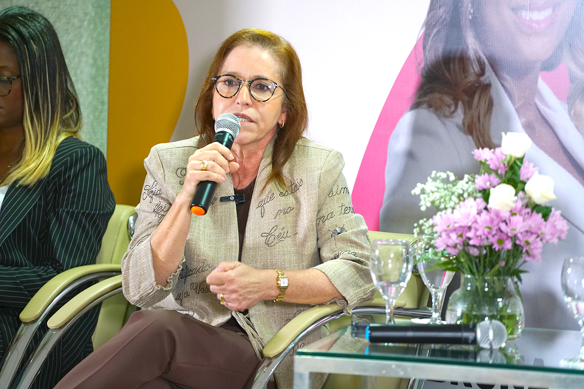 Plano médio de uma mulher de óculos e cabelos castanhos, vestindo um blazer bege com inscrições em caligrafia e calça marrom, falando ao microfone. Ela está sentada em uma poltrona clara, com uma postura firme e expressiva. Em primeiro plano, uma mesa de vidro com um arranjo de flores rosas e brancas; ao fundo, parte de um painel com elementos em rosa e amarelo.