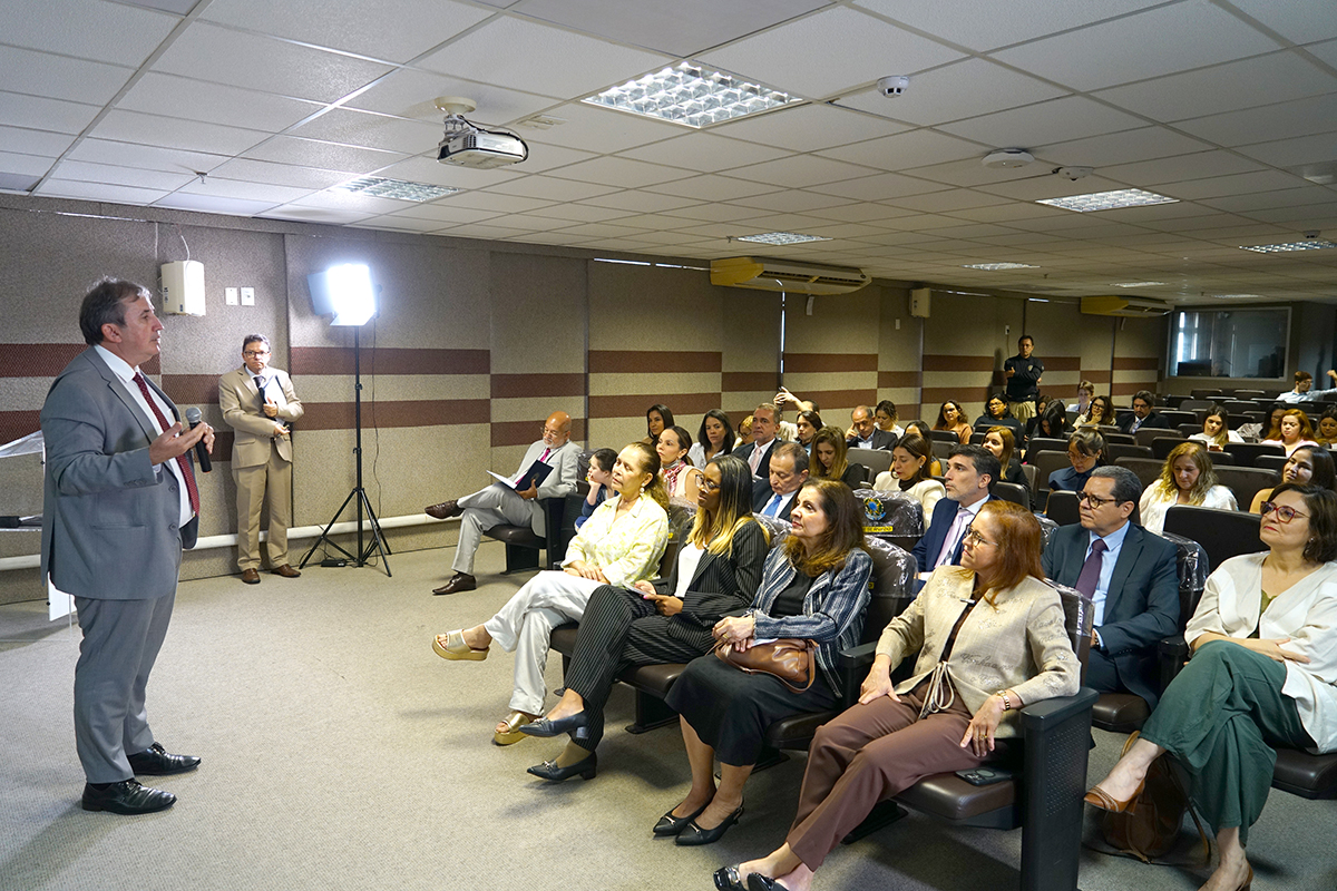 Fotografia em plano aberto de um auditório durante uma palestra. À esquerda, um homem de terno cinza e gravata vermelha fala ao microfone, de frente para a plateia. O público, composto majoritariamente por mulheres e homens em trajes formais, ocupa fileiras de poltronas escuras e acompanha a fala com atenção. O ambiente possui iluminação de teto e um refletor profissional ao fundo, reforçando o caráter institucional do evento.