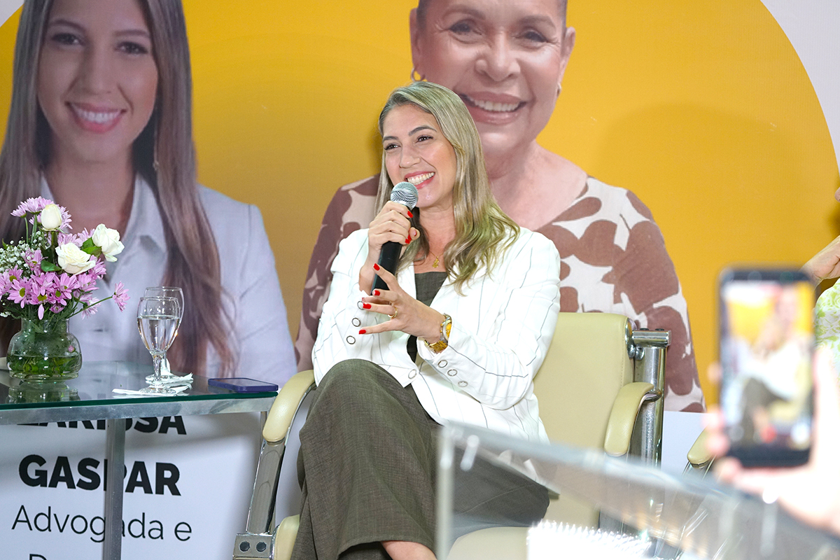 Plano médio de uma mulher loira e sorridente, sentada em uma poltrona bege, falando ao microfone. Ela veste um blazer branco com listras finas e calça escura, destacando-se pelas unhas pintadas de vermelho. Ao fundo, um painel amarelo exibe fotos de outras mulheres, e à esquerda há uma mesa de vidro com flores e um copo d'água. No canto inferior direito, um celular desfocado registra a cena.
