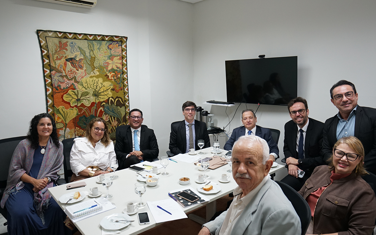 Fotografia colorida em plano médio de nove pessoas sentadas ao redor de uma mesa de reunião branca. Na mesa há copos d'água, xícaras de café, pratos com biscoitos e pães, celulares, papéis e canetas. No fundo, há uma parede branca com uma tapeçaria colorida com motivos de natureza e uma televisão preta.