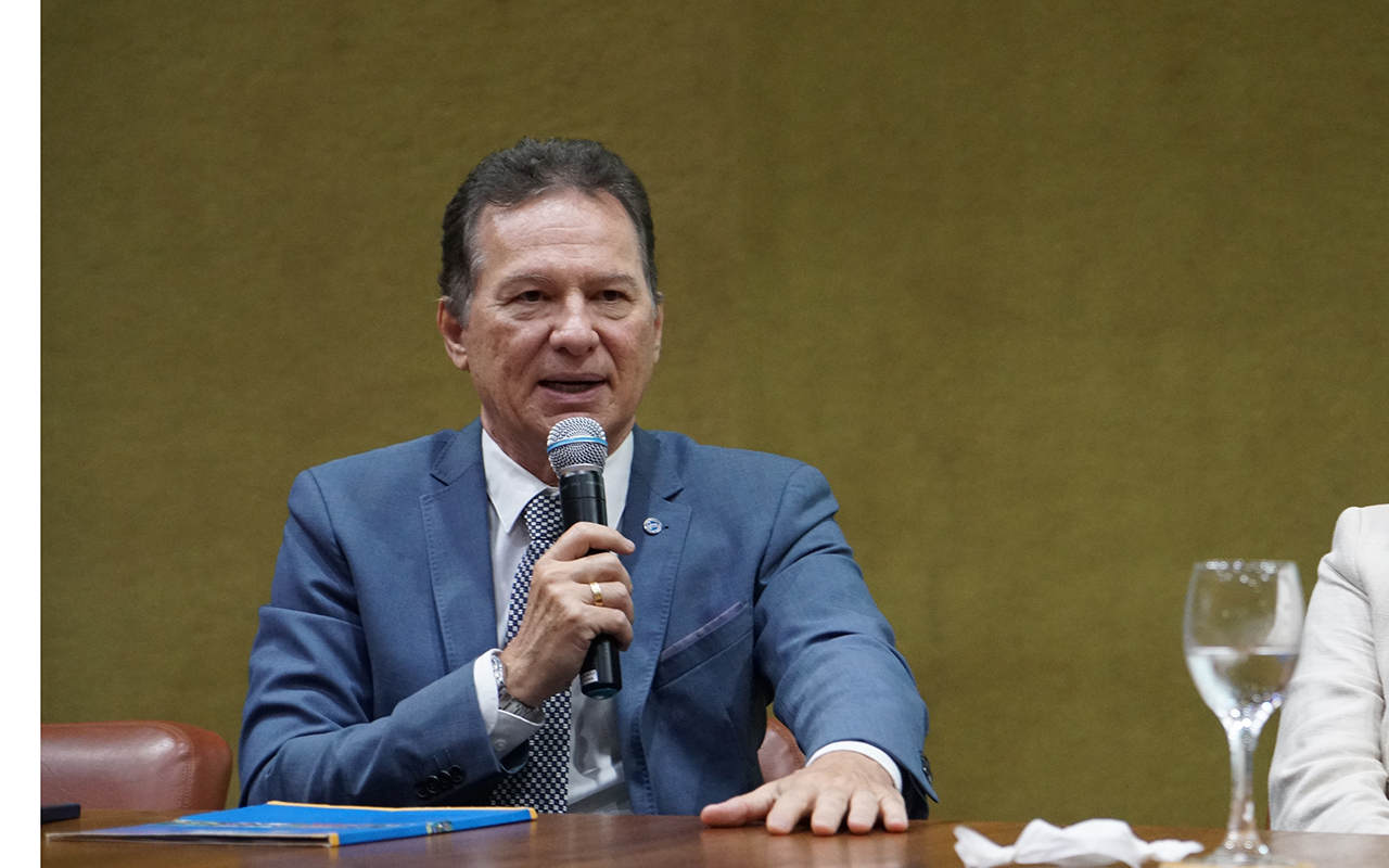 Uma fotografia de plano médio captura um homem de meia-idade, com cabelos castanhos curtos e grisalhos, vestindo um terno azul-petróleo, camisa branca e uma gravata de bolinhas escuras. Ele está sentado atrás de uma mesa de madeira polida, segurando um microfone sem fio na mão direita, próximo à boca, enquanto a mão esquerda repousa sobre a mesa. Sua expressão é focada e engajada, como se estivesse discursando ou respondendo a uma pergunta em um painel de discussão. O fundo é uma parede texturizada de cor verde-oliva, com um toque de uma pessoa em trajes brancos e um copo de água visível no lado direito da moldura.