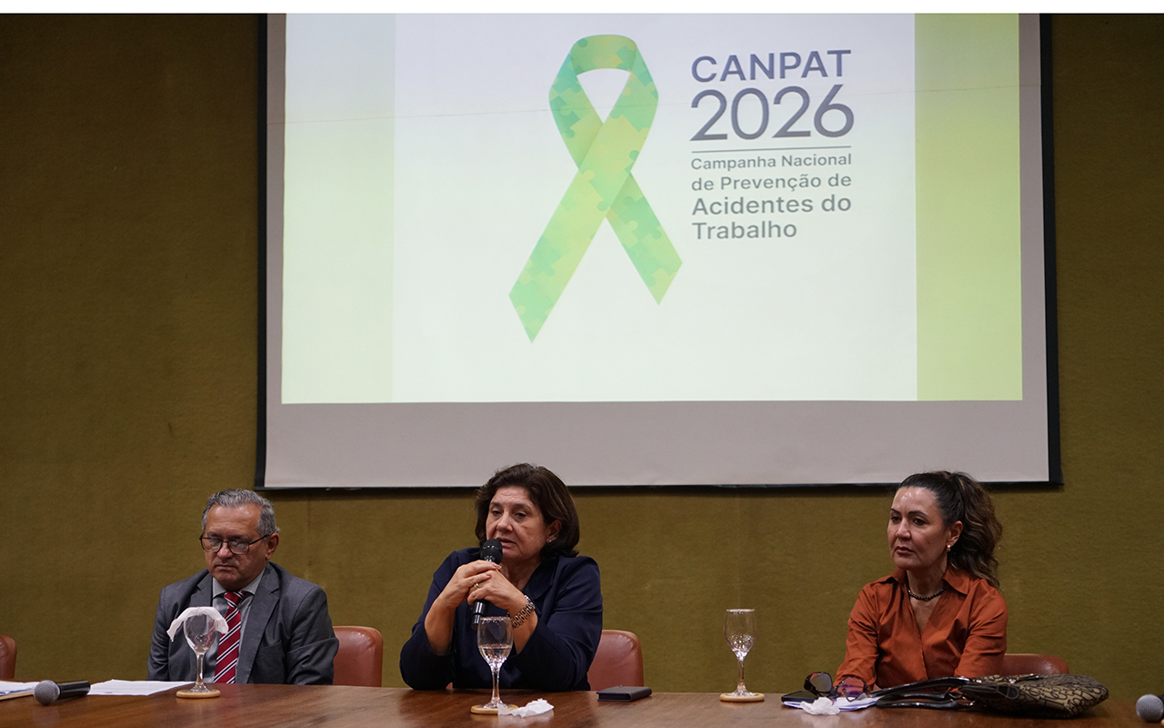 Três pessoas estão sentadas à mesa de uma comissão diretora em um auditório, com uma tela de projeção ao fundo exibindo o logotipo da "CANPAT 2026", um laço verde de quebra-cabeça e o texto "Campanha Nacional de Prevenção de Acidentes do Trabalho". A oradora principal, uma mulher de cabelos curtos e jaqueta escura no centro, fala ao microfone, ladeada por um homem de terno cinza e gravata vermelha à esquerda e uma mulher de camisa cor de ferrugem à direita. Sobre a mesa de madeira há taças de água, microfones, papéis e uma bolsa.