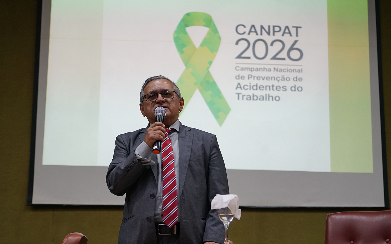 Um homem de cabelos grisalhos e óculos, vestindo paletó cinza e gravata listrada em tons de vermelho e branco, fala ao microfone em um auditório. Ele está posicionado à frente de uma grande tela de projeção que exibe o logotipo da "CANPAT 2026" com o laço verde e a frase "Campanha Nacional de Prevenção de Acidentes do Trabalho". A imagem é capturada de um ângulo levemente de baixo para cima, destacando o orador contra o fundo institucional da campanha.