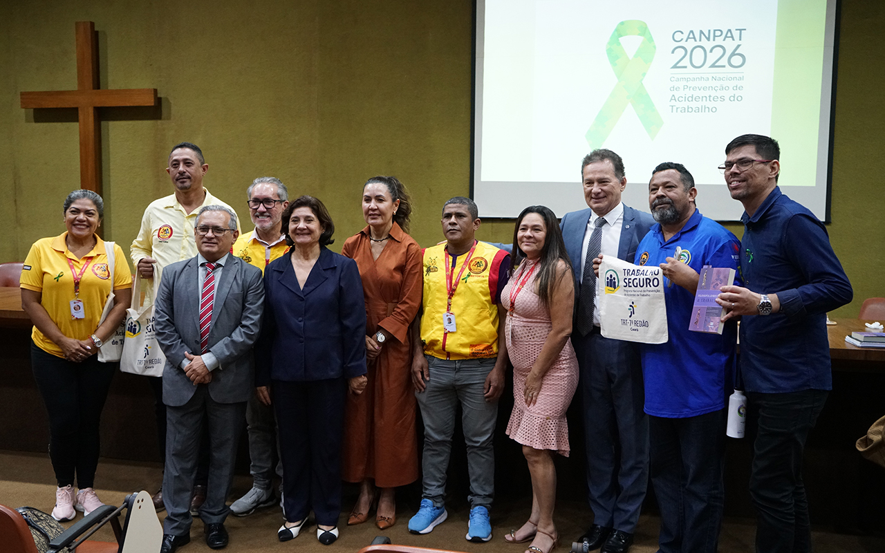 Uma foto de grupo com dez pessoas posando em um auditório em frente a uma tela de projeção. No centro do palco, as pessoas vestem roupas profissionais e uniformes temáticos, como camisas amarelas e azuis com laços verdes, símbolos da campanha CANPAT 2026. A tela ao fundo exibe o logotipo "CANPAT 2026", um laço verde de quebra-cabeça e o texto "Campanha Nacional de Prevenção de Acidentes do Trabalho", tudo com um grande crucifixo de madeira à esquerda.