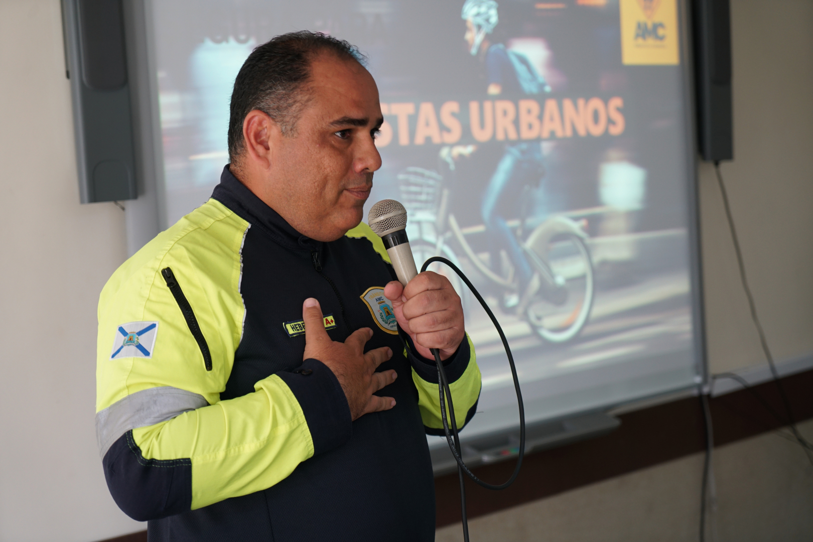 Homem de pele parda e cabelos curtos escuros, vestindo o uniforme da AMC em cores preto e amarelo fluorescente, fala ao microfone segurando-o com a mão esquerda. Ele está de perfil para a direita, com a mão direita sobre o peito, em frente a uma tela de projeção que exibe a imagem de um ciclista e o texto "Ciclistas Urbanos". O palestrante demonstra expressão serena e engajada durante a oficina de pilotagem segura.