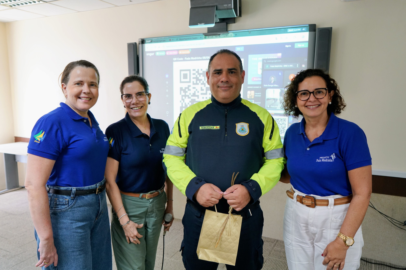 O agente de segurança Heberfran Bruno, ao centro e segurando uma sacola de presente dourada, posa para foto ao lado de três servidoras do subcomitê do Programa Fada Madrinha. Elas vestem camisas polo azuis com o logotipo do projeto, enquanto o agente utiliza seu uniforme preto e amarelo da AMC. O grupo sorri em frente a uma tela digital que exibe um QR Code na sala de aula da Escola Judicial.