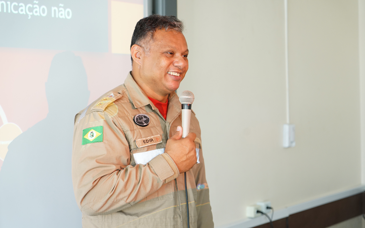 Fotografia colorida, em plano médio, de um bombeiro militar sorrindo e falando ao microfone. Ele é um homem de pele morena e cabelos grisalhos curtos, usando uniforme operacional cáqui com a bandeira do Ceará na manga e a identificação "EDIR". O bombeiro está em um ambiente interno, com uma parede branca à direita e uma tela de projeção à esquerda, onde se vê o final da frase "nicação não" e sua sombra refletida.