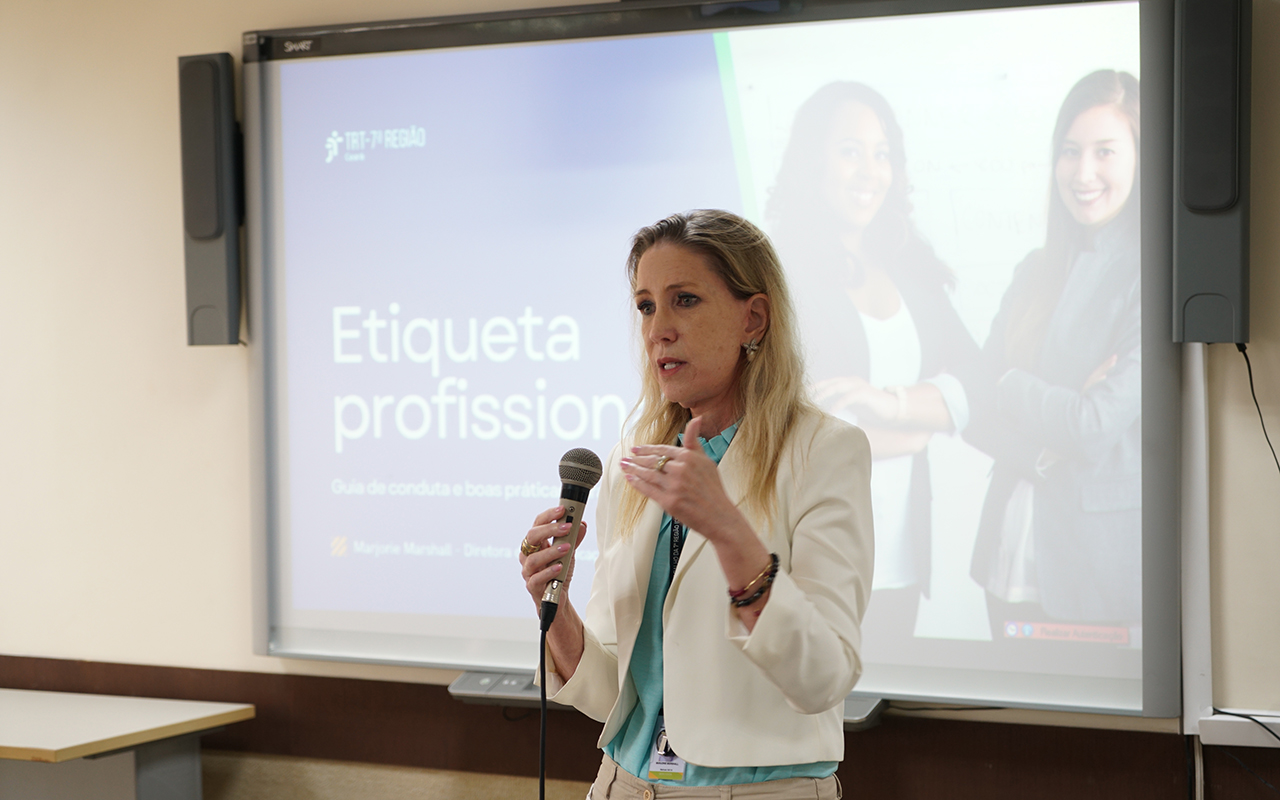 Fotografia colorida, em plano médio, de uma mulher loira de meia-idade palestrando em uma sala de aula. Ela veste um blazer branco sobre uma blusa azul-clara, calças cáqui e usa crachá. Segurando um microfone na mão direita e gesticulando com a esquerda, ela fala em frente a uma lousa digital SMART Board. A tela exibe a apresentação "Etiqueta profissional: Guia de conduta e boas práticas", com a logomarca do TRT 7ª Região no canto superior esquerdo e a foto de duas mulheres profissionais sorrindo à direita.