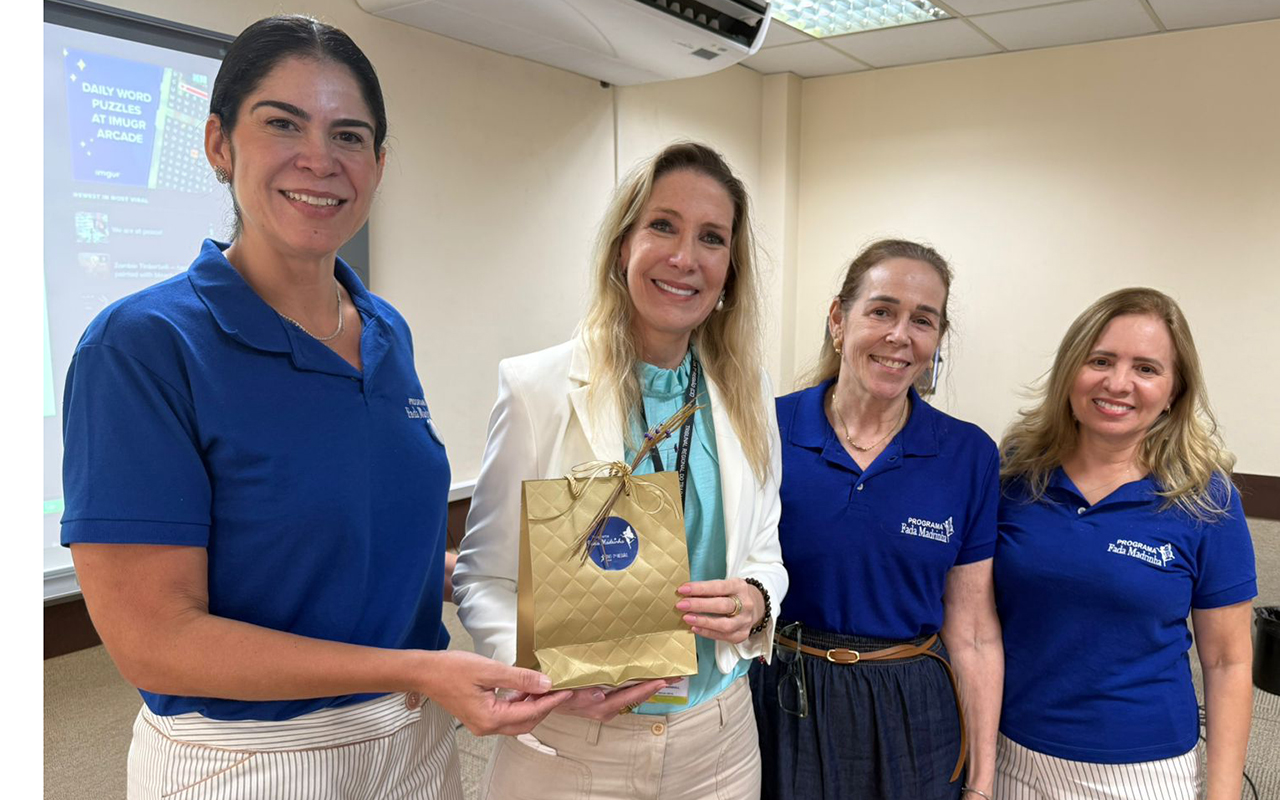 Fotografia colorida em plano médio de quatro mulheres sorrindo em uma sala de aula. À esquerda, a juíza Ana Caroline veste camisa polo azul com a logo do Programa Fada Madrinha e entrega uma sacola de presente dourada à palestrante Marjorie Marshall, que veste blazer branco. À direita, as servidoras Sylvianne Fontenelle e Rejane Facanha, ambas vestindo a polo azul do programa, completam o registro da entrega da lembrança.