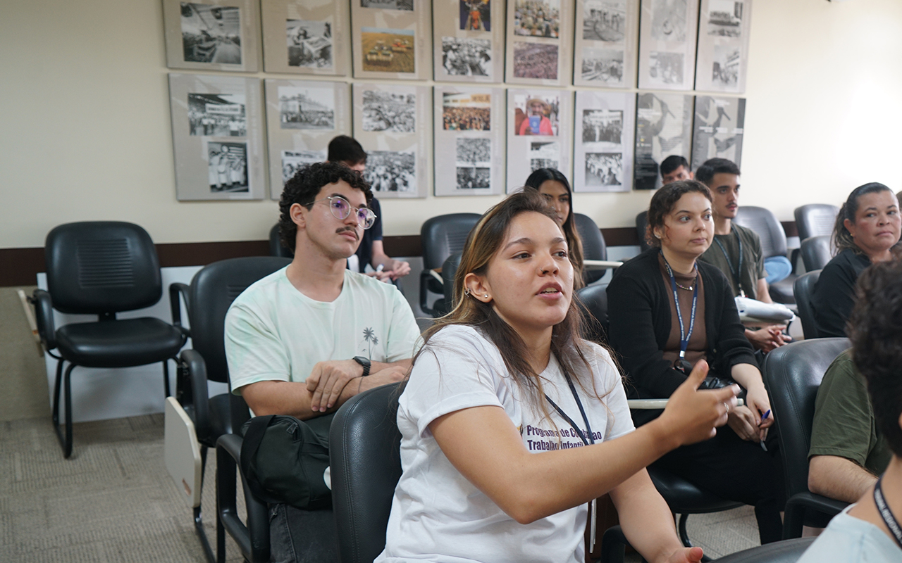 Em primeiro plano, uma jovem de camisa branca com a logo de combate ao trabalho infantil fala e gesticula com a mão direita em um auditório. Atrás dela, outros jovens e adultos assistem atentamente à apresentação, sentados em cadeiras de estofado preto com pranchetas acopladas. Na parede ao fundo, observa-se uma galeria de fotos emolduradas em preto e branco.