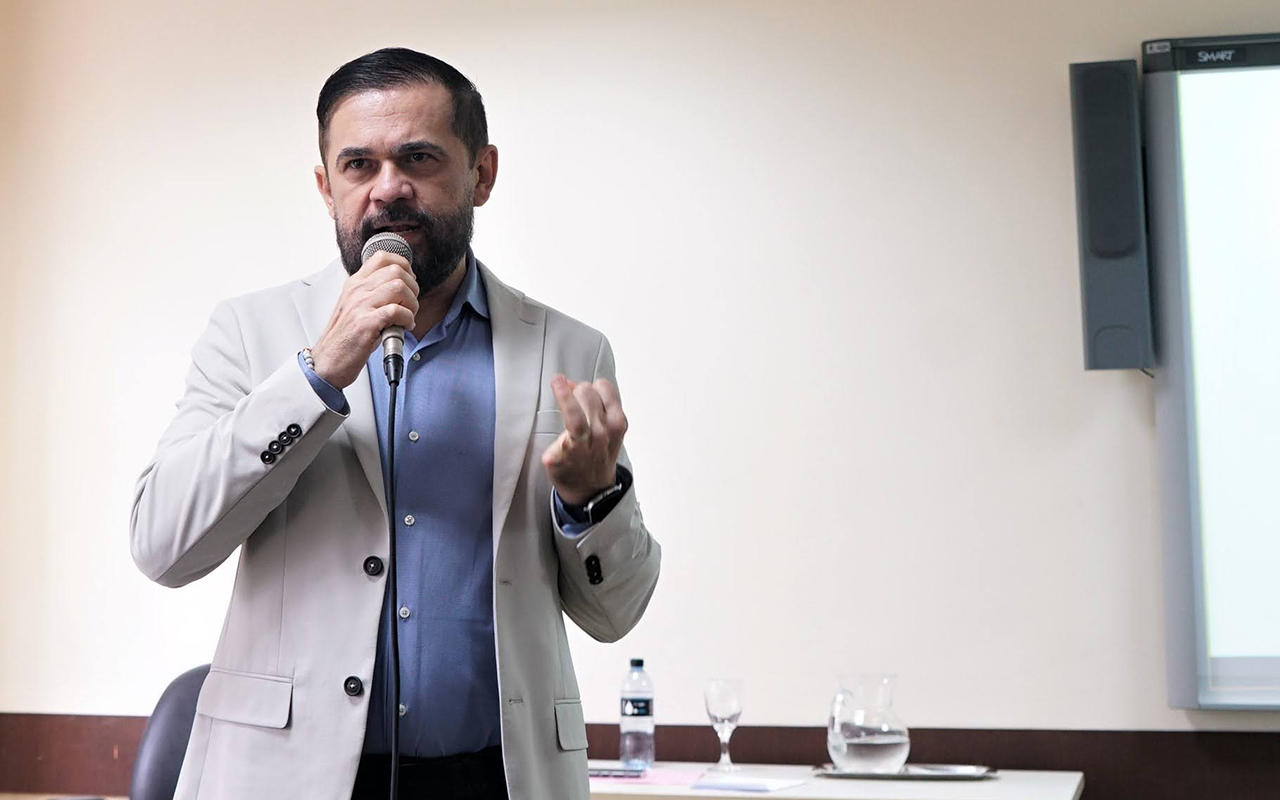 Um homem branco de barba aparada, vestindo paletó bege sobre camisa azul, fala ao microfone enquanto faz um gesto com a mão esquerda. Ele está em um auditório, e ao fundo observa-se uma mesa com água e parte de uma tela de projeção.