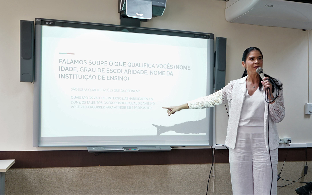 Uma mulher branca, de cabelos pretos presos, veste terno branco e fala ao microfone. Ela aponta para uma lousa digital que exibe um slide sobre qualificações e valores internos. Ao fundo, uma parede clara com equipamentos de som e ar-condicionado.