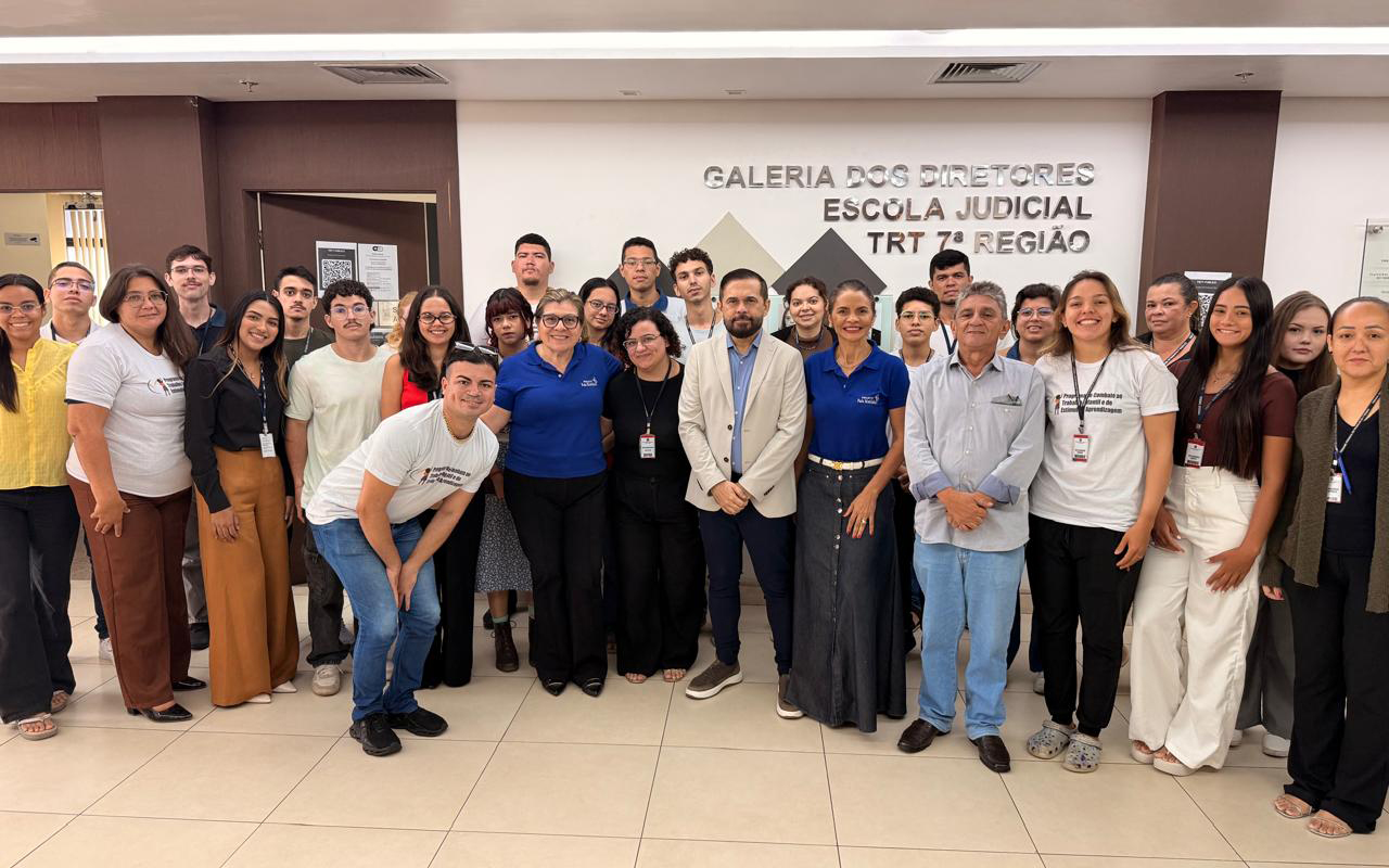 Um grupo de cerca de trinta pessoas posa sorridente em um hall amplo. Ao centro, destacam-se os juízes Célio Timbó e Ana Caroline, ladeados por estagiários e servidores, alguns vestindo a camisa branca do Programa de Combate ao Trabalho Infantil. Ao fundo, a inscrição "Galeria dos Diretores - Escola Judicial TRT 7ª Região".