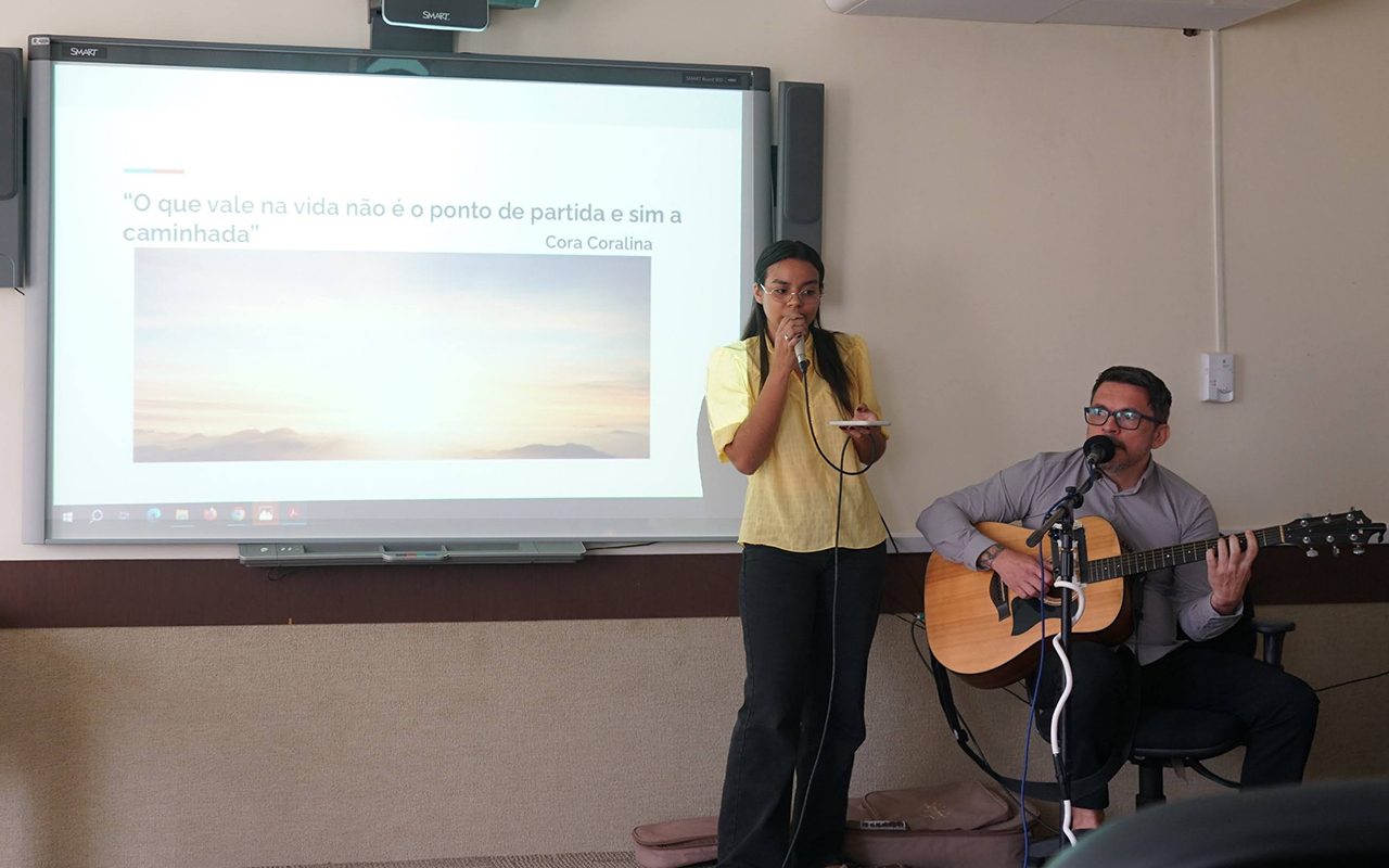 Uma jovem de blusa amarela canta ao microfone ao lado de um homem sentado que toca violão. Eles estão à frente de uma tela de projeção que exibe uma frase de Cora Coralina sobre um fundo de paisagem ao pôr do sol. A cena ocorre em um auditório com paredes claras e acabamento em madeira escura.