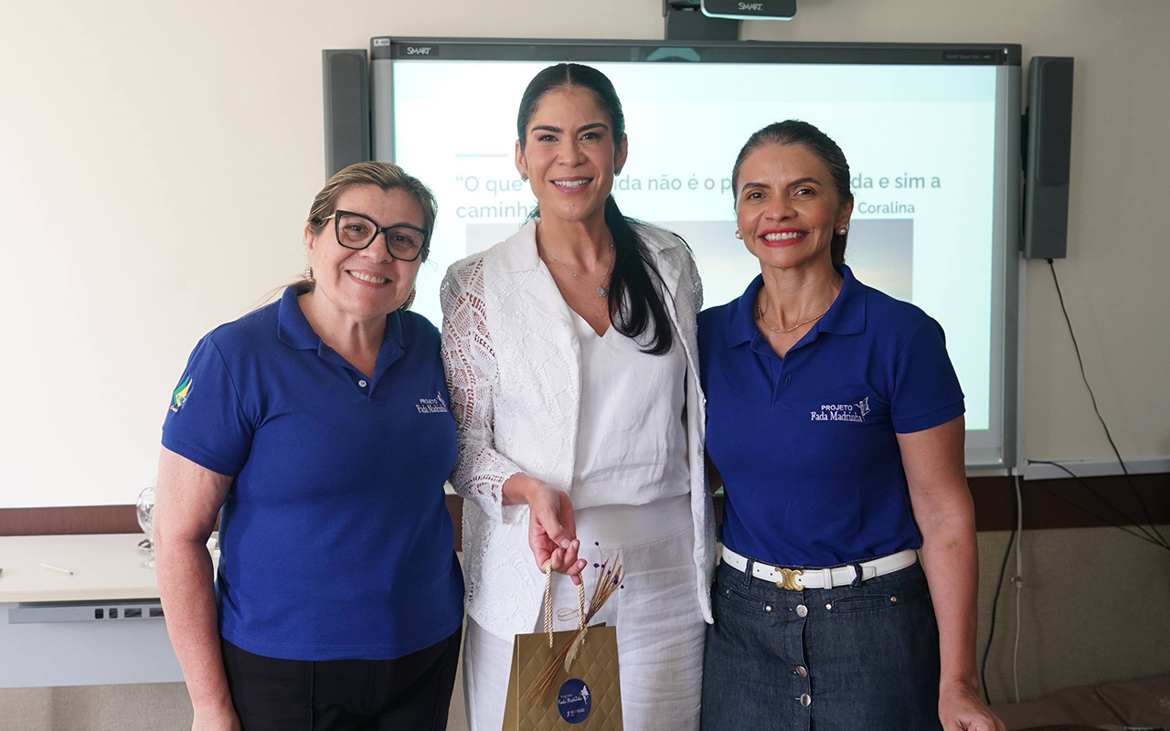 Três mulheres posam sorridentes para a foto em frente à lousa digital que projeta o slide com a frase de Cora Coralina. A mulher ao centro, de terno branco, recebe uma sacola de presente dourada das outras duas mulheres que vestem camisas polo azuis com a logo do "Projeto Fada Madrinha". A sala de aula possui equipamentos de áudio e vídeo e uma mesa branca ao fundo.