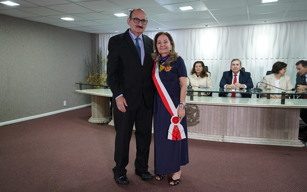 Um homem de terno escuro e uma mulher posam em pé para a foto. Ela usa um vestido longo azul-marinho e uma faixa transversal vermelha e branca com a insígnia da medalha. Ao fundo, três autoridades estão sentadas à mesa diretora em um auditório com tons de bege e cinza.