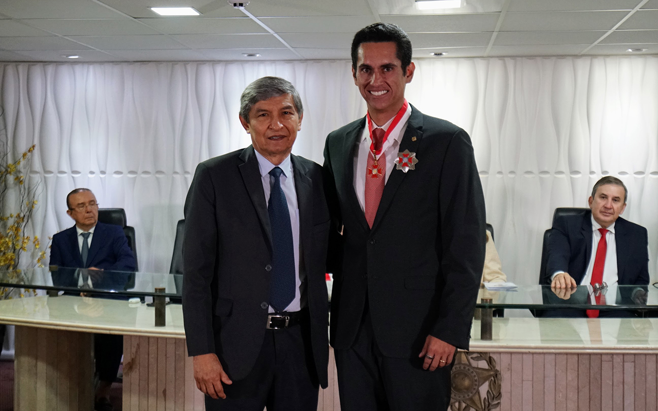Dois homens de terno posam em pé em um salão de eventos, o da direita com uma medalha vermelha ao pescoço. Atrás deles, uma mesa longa de vidro com mais duas autoridades sentadas e, ao fundo, uma parede texturizada de painéis brancos. No teto, luminárias quadradas embutidas e piso de carpete.