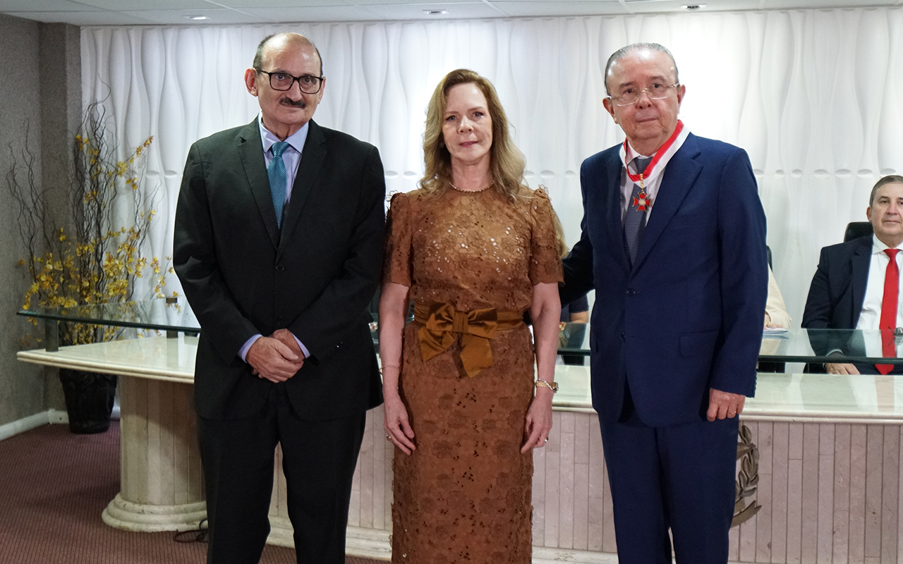 Uma mulher com vestido de renda marrom e laço na cintura posa entre dois homens de terno. O homem à direita veste terno azul e exibe a medalha com fita vermelha no pescoço; o da esquerda usa terno preto e óculos. Ao fundo, vê-se parte da mesa diretora e uma parede branca com relevos ondulados.