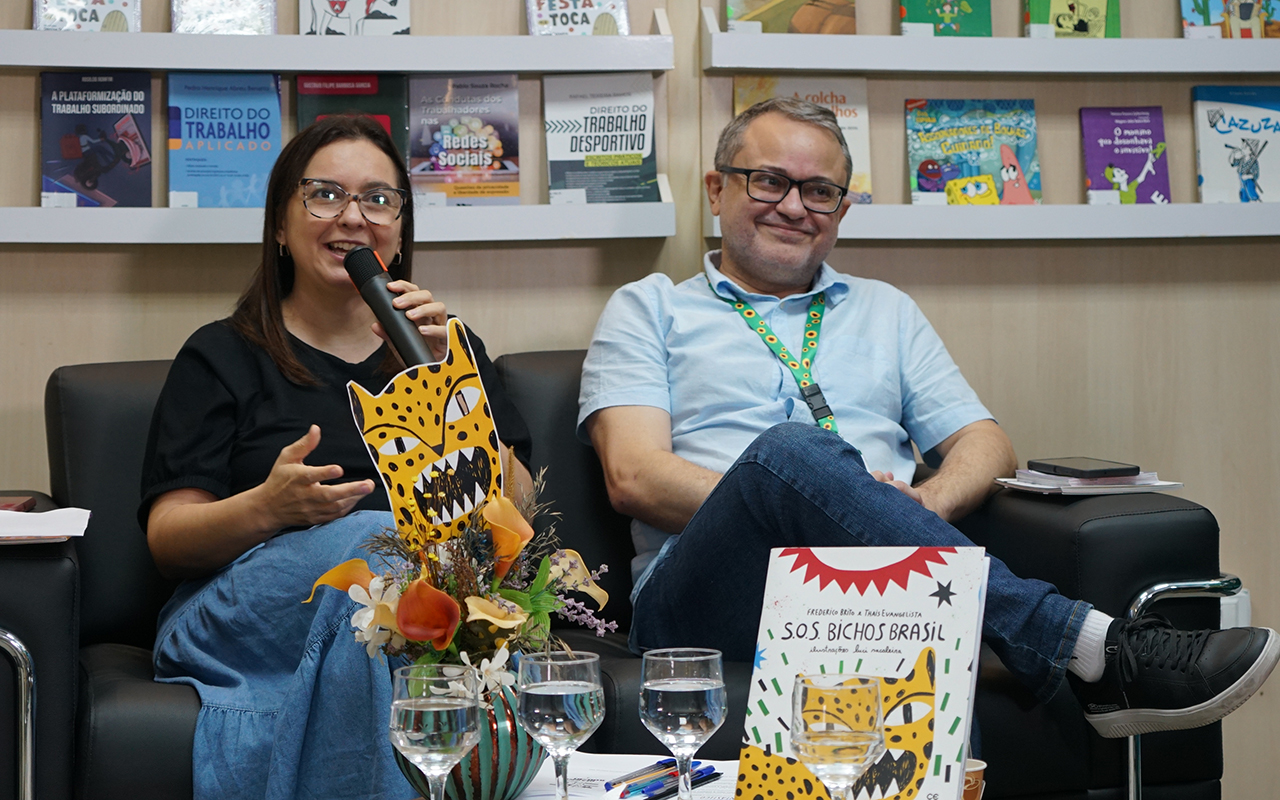 Uma mulher com óculos e camisa preta fala ao microfone e faz gestos com a mão esquerda, sentada em um sofá preto ao lado de um homem sorridente com óculos e camisa azul claro, em um ambiente de biblioteca. No centro, entre os dois, há um vaso de flores com recortes de animais (onça) e à frente, sobre uma mesa, dois copos com água, canetas e o livro "S.O.S. Bichos Brasil". Ao fundo, estantes de madeira branca expõem diversos livros com capas coloridas.