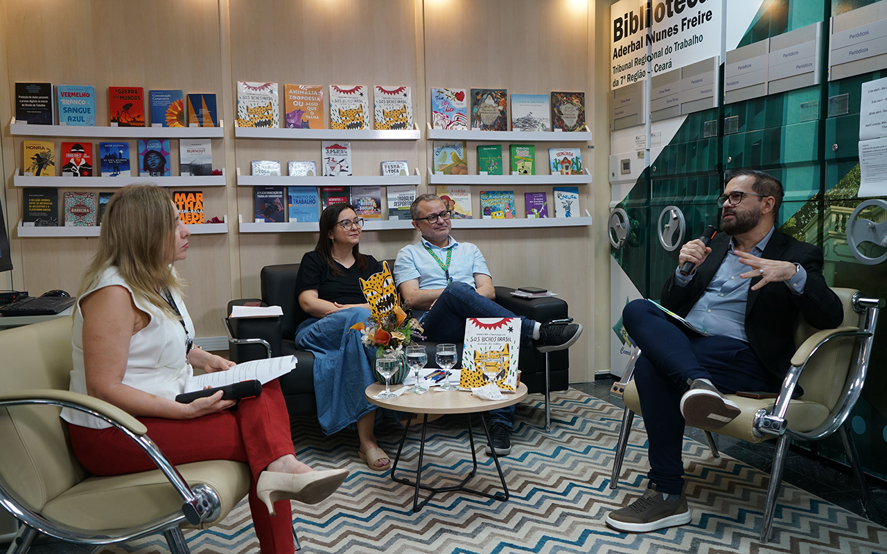 Em um plano aberto, quatro pessoas participam de um bate-papo em uma biblioteca com estantes repletas de livros ao fundo. À direita, um homem de terno fala ao microfone, enquanto ao centro um casal no sofá e uma mulher à esquerda, em uma poltrona, acompanham a conversa. O ambiente é decorado com um tapete geométrico em tons de bege e azul, e sobre a mesa central destaca-se o livro "S.O.S. Bichos Brasil".